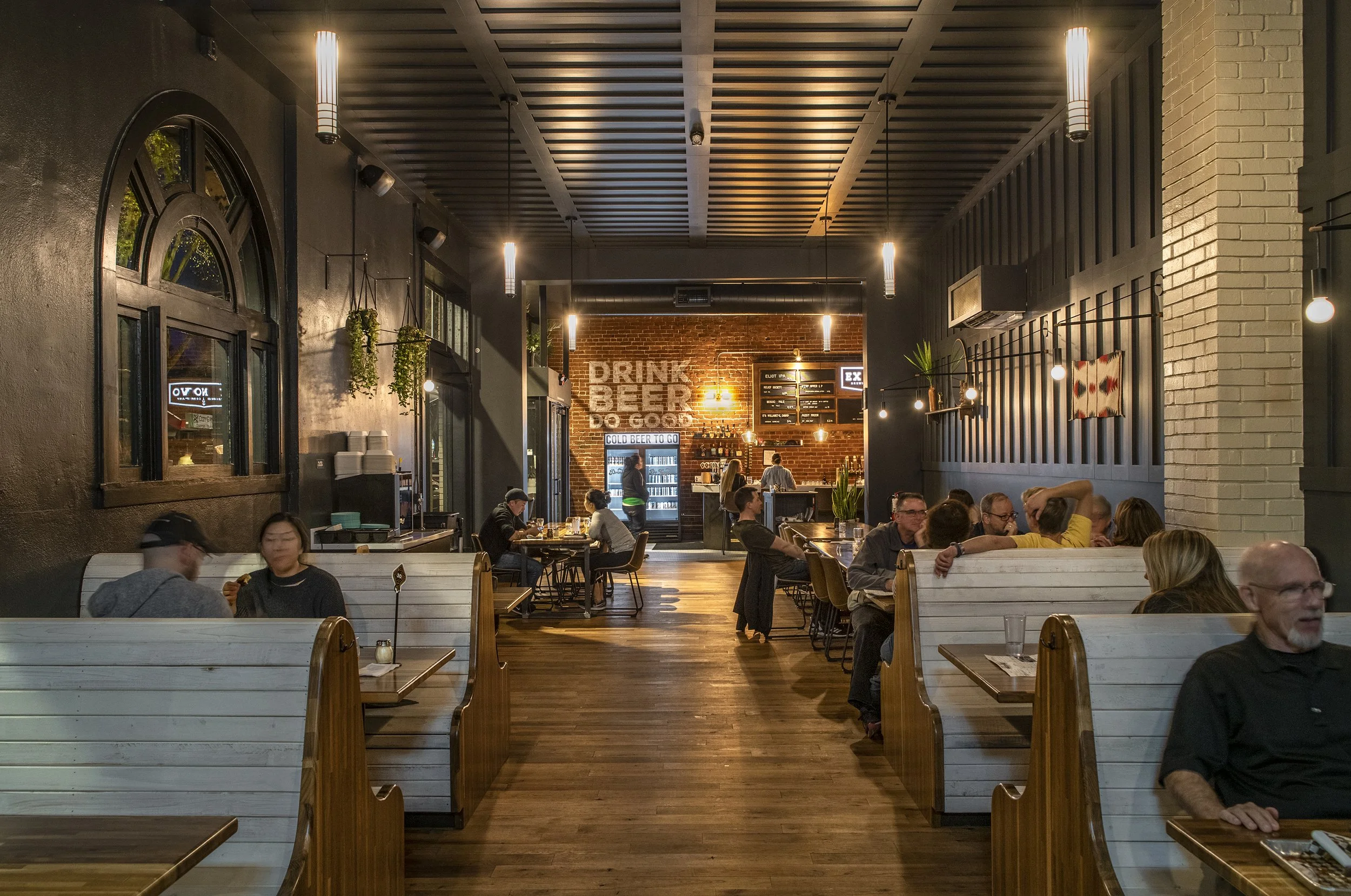 Interior of a modern restaurant with white booth seating, dark walls, hanging lights, and a brick wall with a drink menu in the background. Customers are dining and socializing.