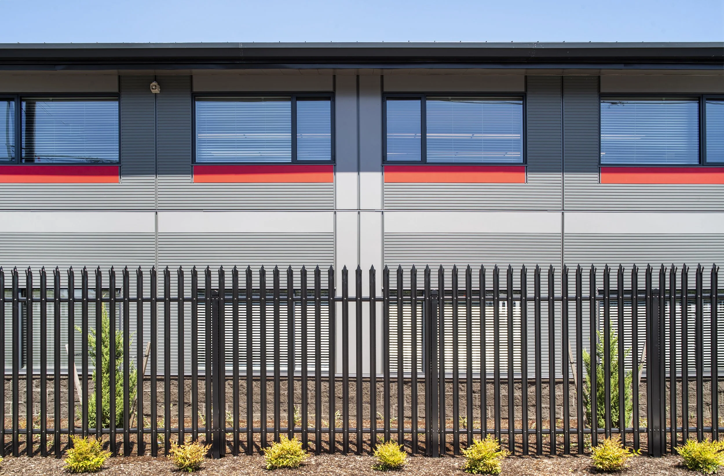 Modern building with grey and black exterior, red accents, large windows with blinds, surrounded by black metal fence and landscaped bushes.