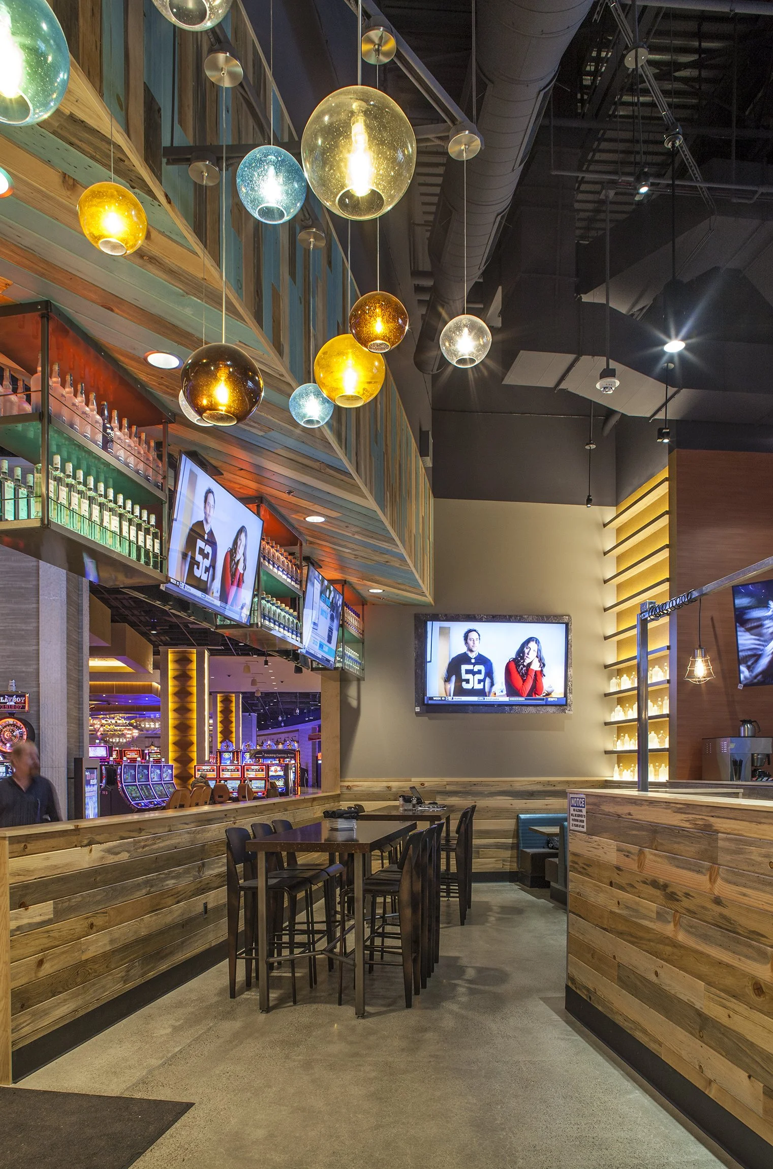 Interior of a modern bar or restaurant with wooden paneling, high tables and chairs, multiple flat-screen TVs showing sports or news, amber, turquoise, and clear spherical pendant lights, and a row of liquor bottles on shelves.