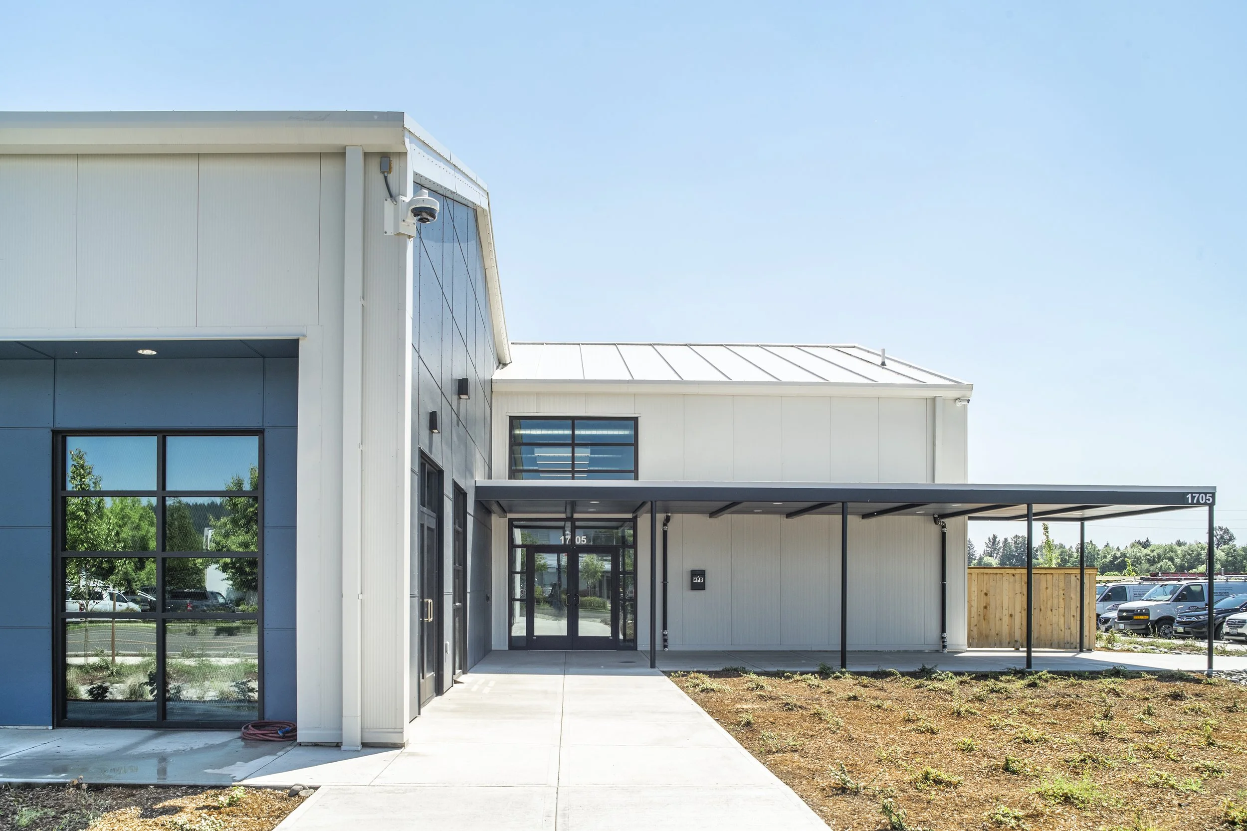 Modern commercial building with black-framed glass doors and windows, covered entrance walkway, and a parking lot in the background under a clear blue sky.