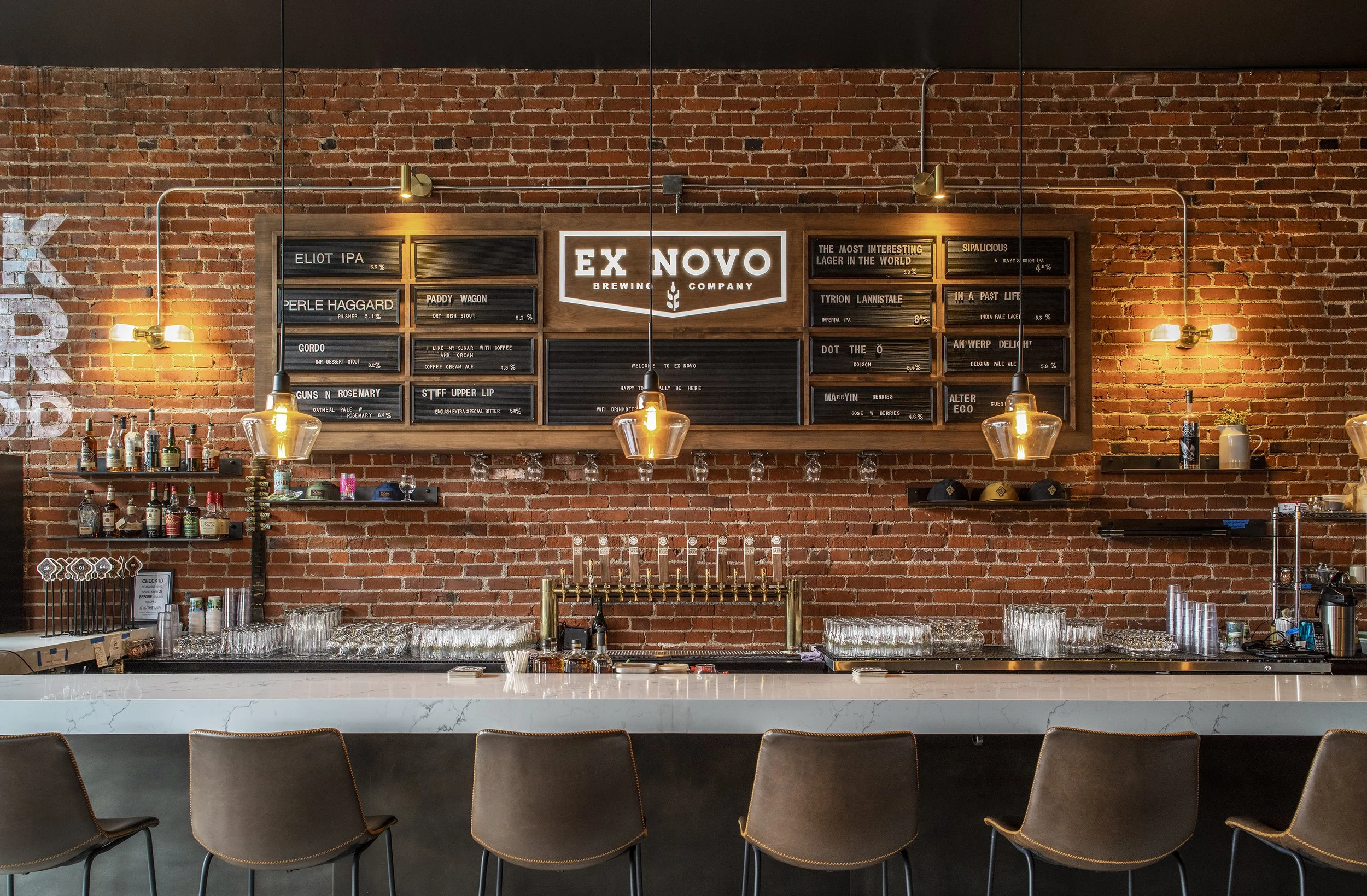Bar with a row of six bar stools in front, a white marble countertop, exposed brick wall with a large chalkboard menu, hanging lights, shelves with bottles and glasses, and beer taps.