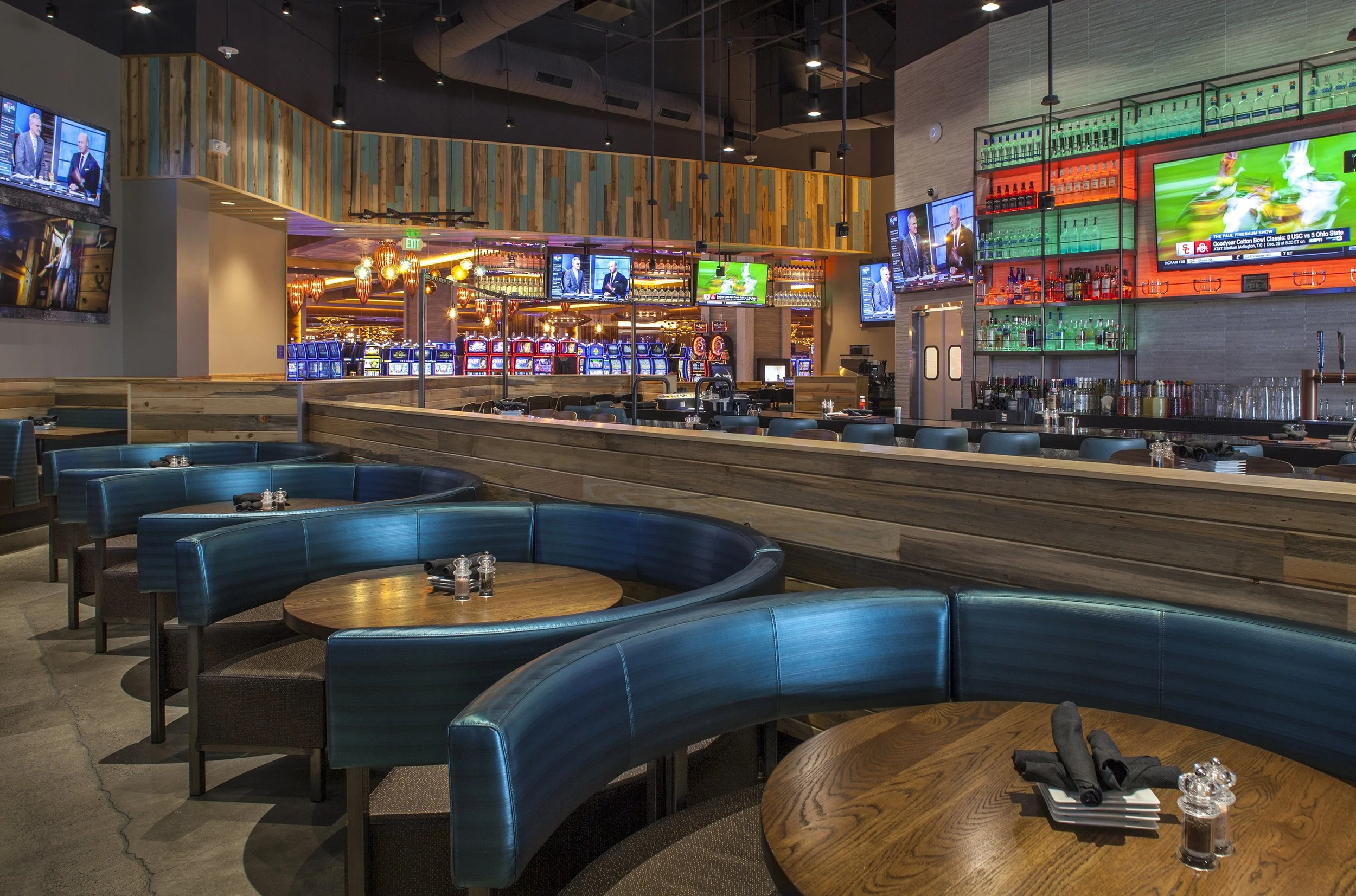 Interior of a modern sports bar or restaurant with round wooden tables, blue cushioned booth seating, multiple flat-screen TVs showing sports and news, a bar area with drinks and glasses, and a slot machine area in the background.