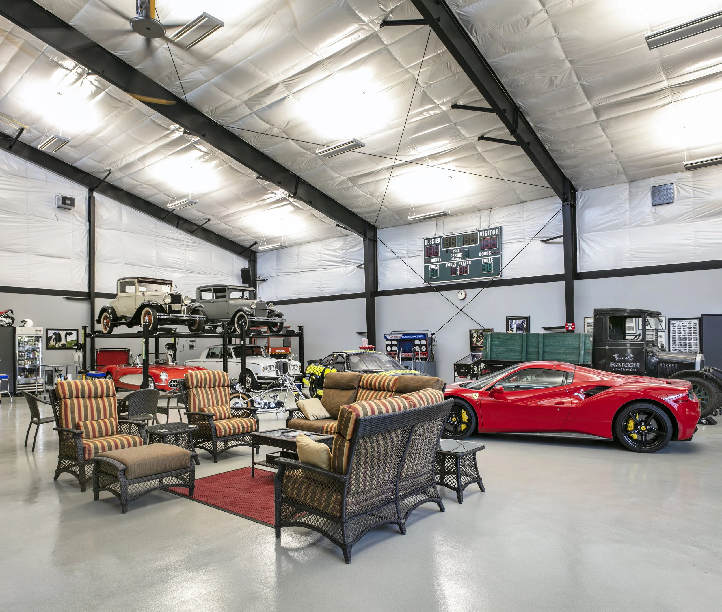 A spacious garage or exhibition space with vintage cars and a modern sports car, including a red Ferrari, surrounded by wicker furniture and vintage arcade machines, a scoreboard, and automotive memorabilia.