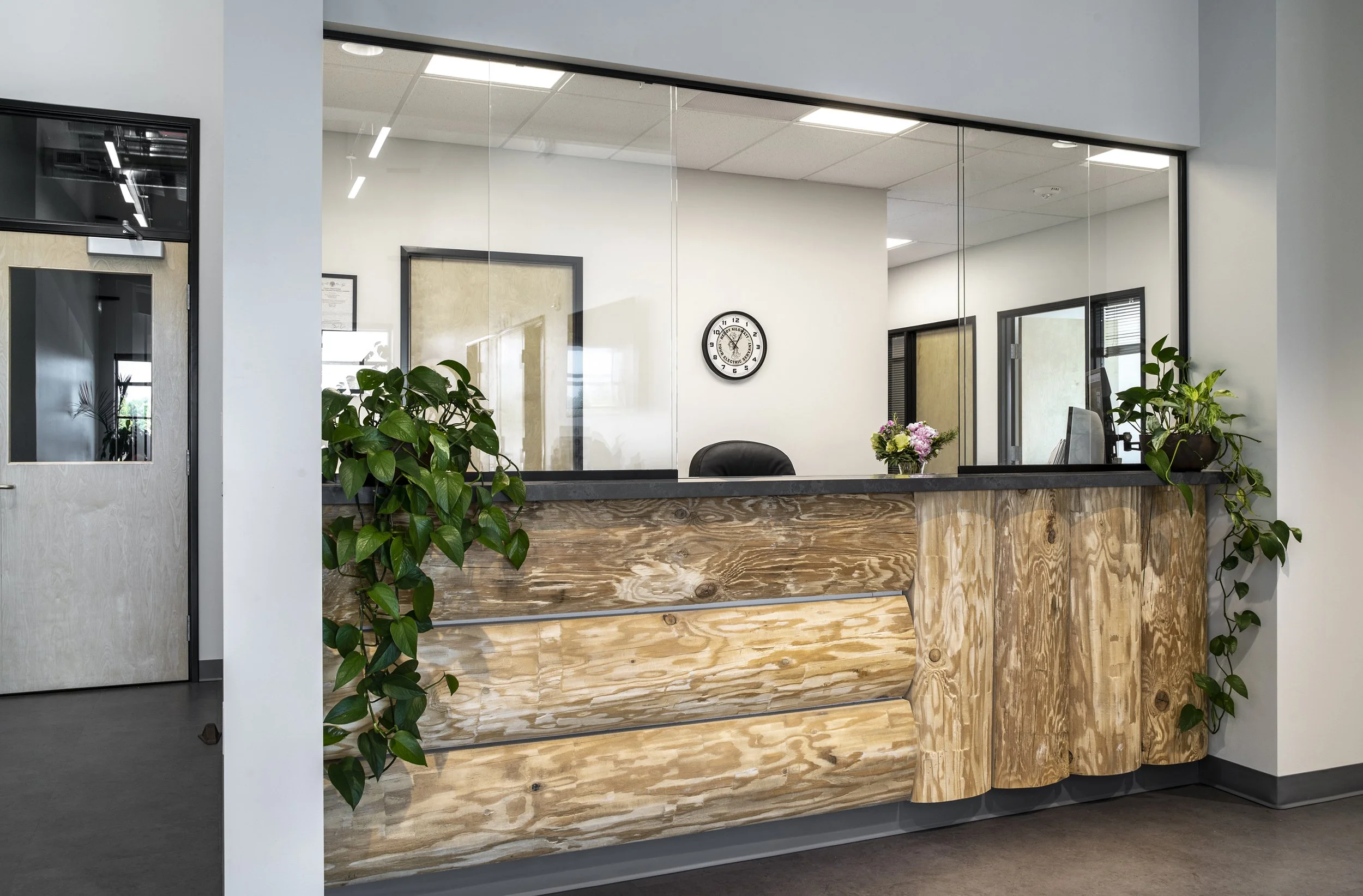 Office reception desk with a glass window behind, decorated with green plants and a small flower arrangement, clock on the wall.