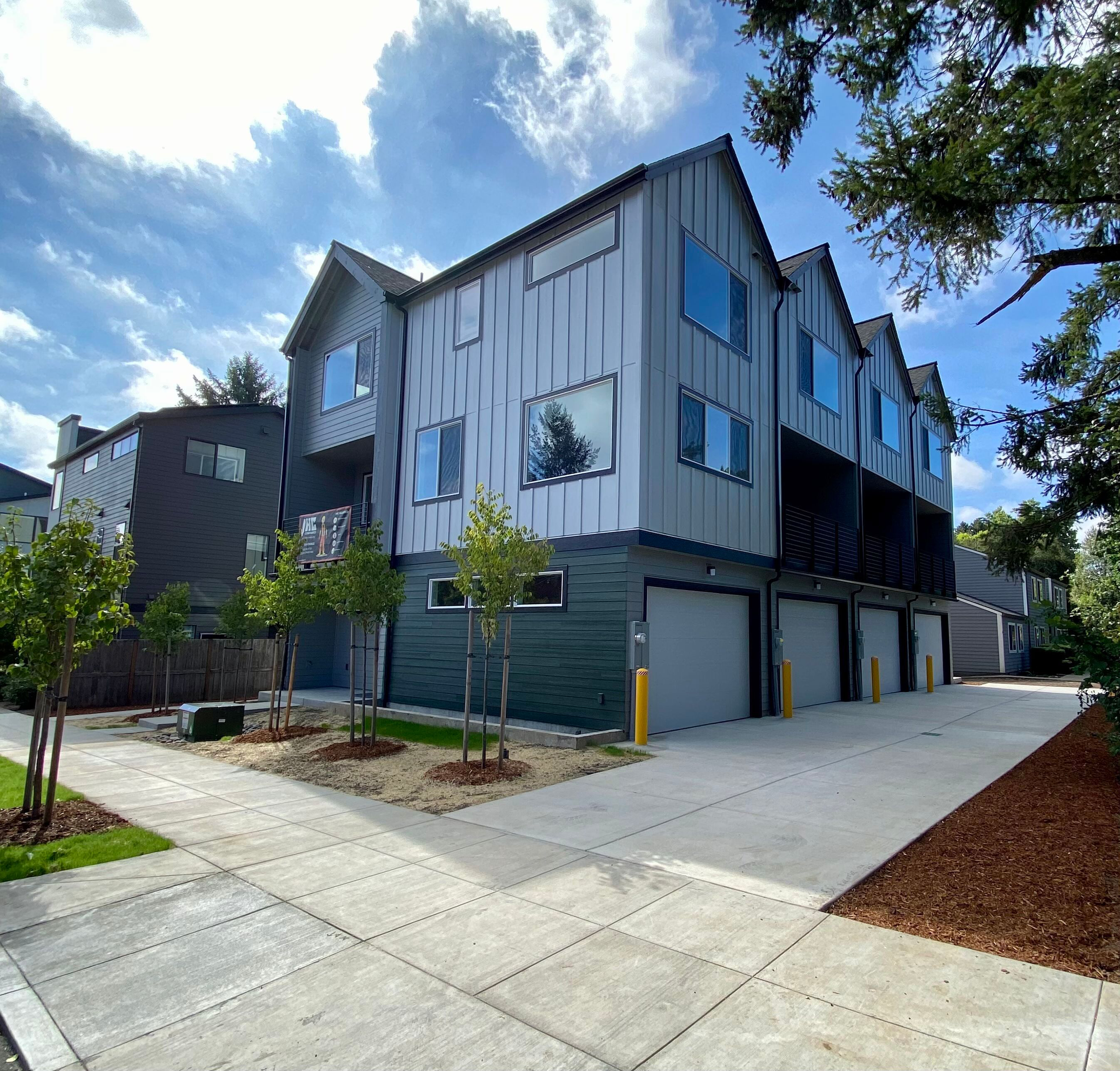 New modern apartment building with four garages, trees, and a sidewalk in front, under a partly cloudy sky.