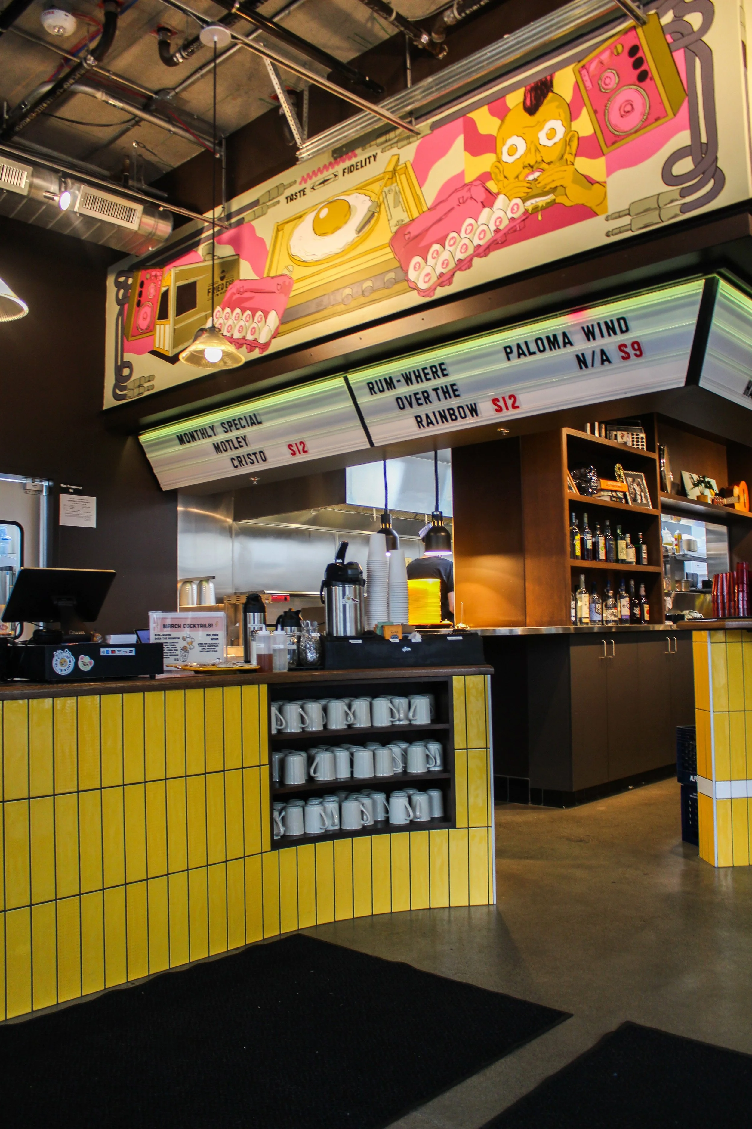 Interior of a cafe or bar with a yellow counters, shelves of mugs and alcohol bottles. A large colorful mural on the wall depicts a person with a buffalo hat and a turntable, with a boombox nearby.
