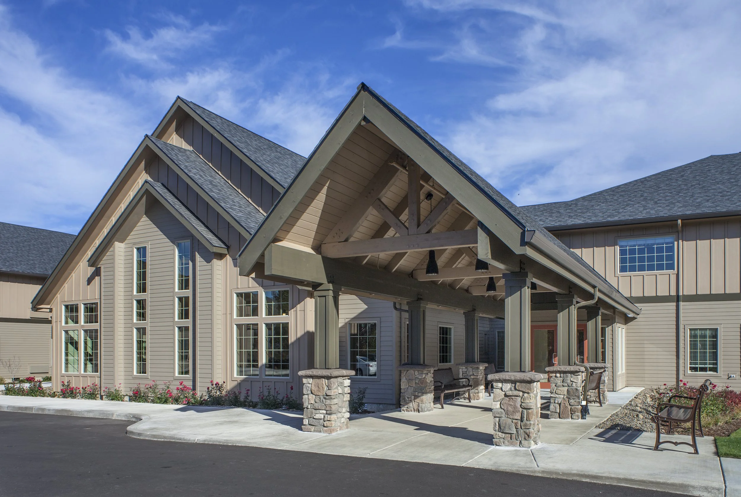 A modern residential building with a covered porch, stone pillars, and multiple large windows, under a partly cloudy blue sky.
