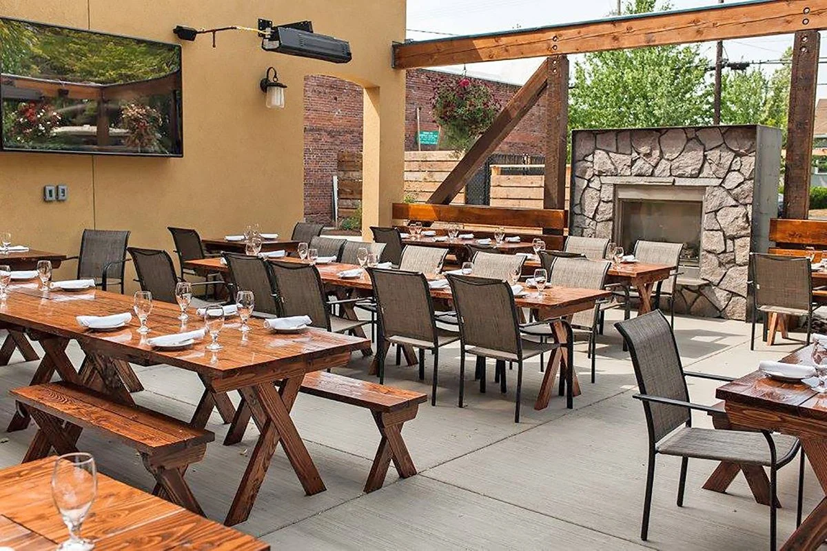 Empty outdoor restaurant patio with wooden tables, chairs, and picnic benches, set with glasses and plates, with a stone fireplace and string lights.