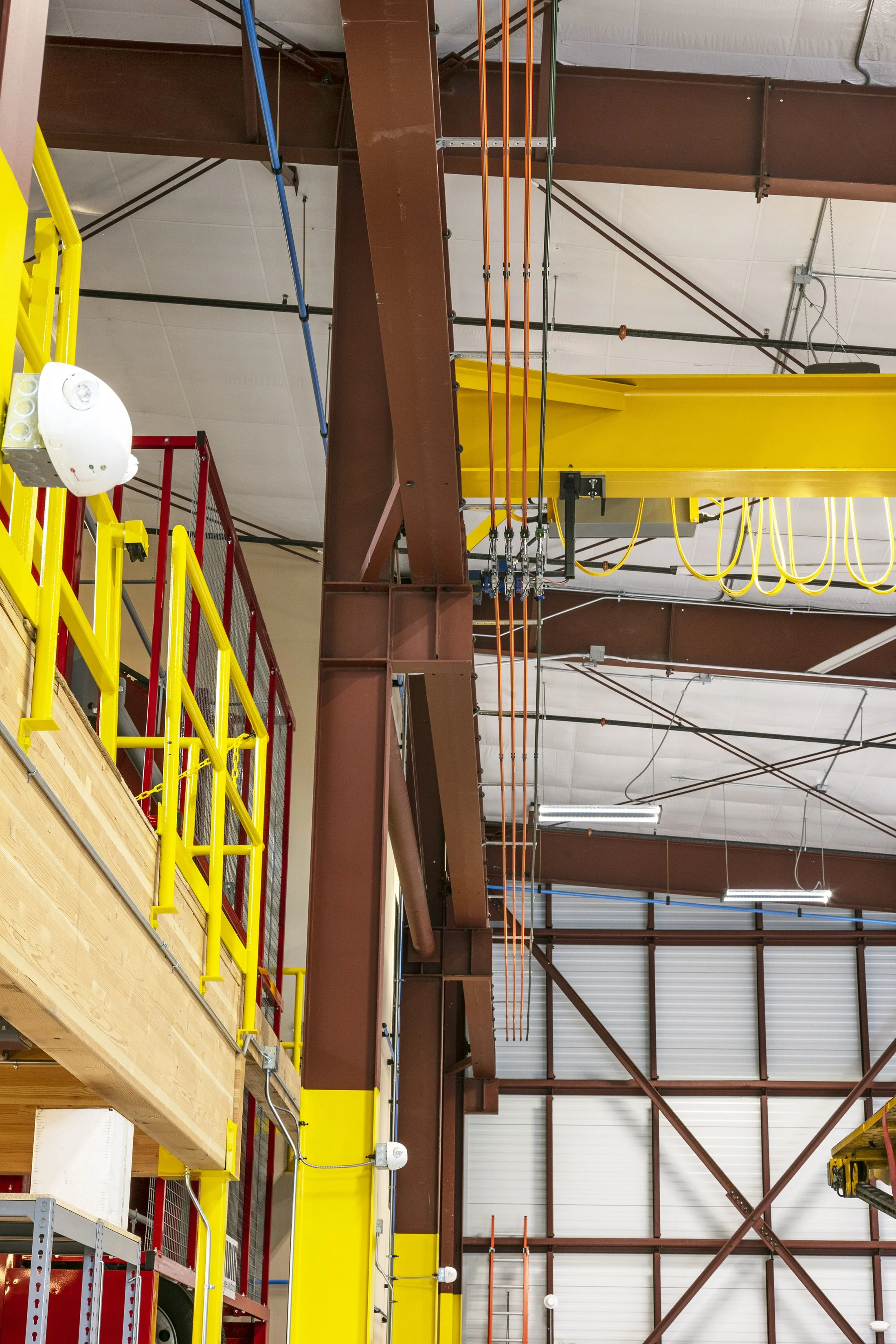 Interior of an industrial warehouse with steel beams, yellow safety rails, and a yellow overhead crane with orange hooks.