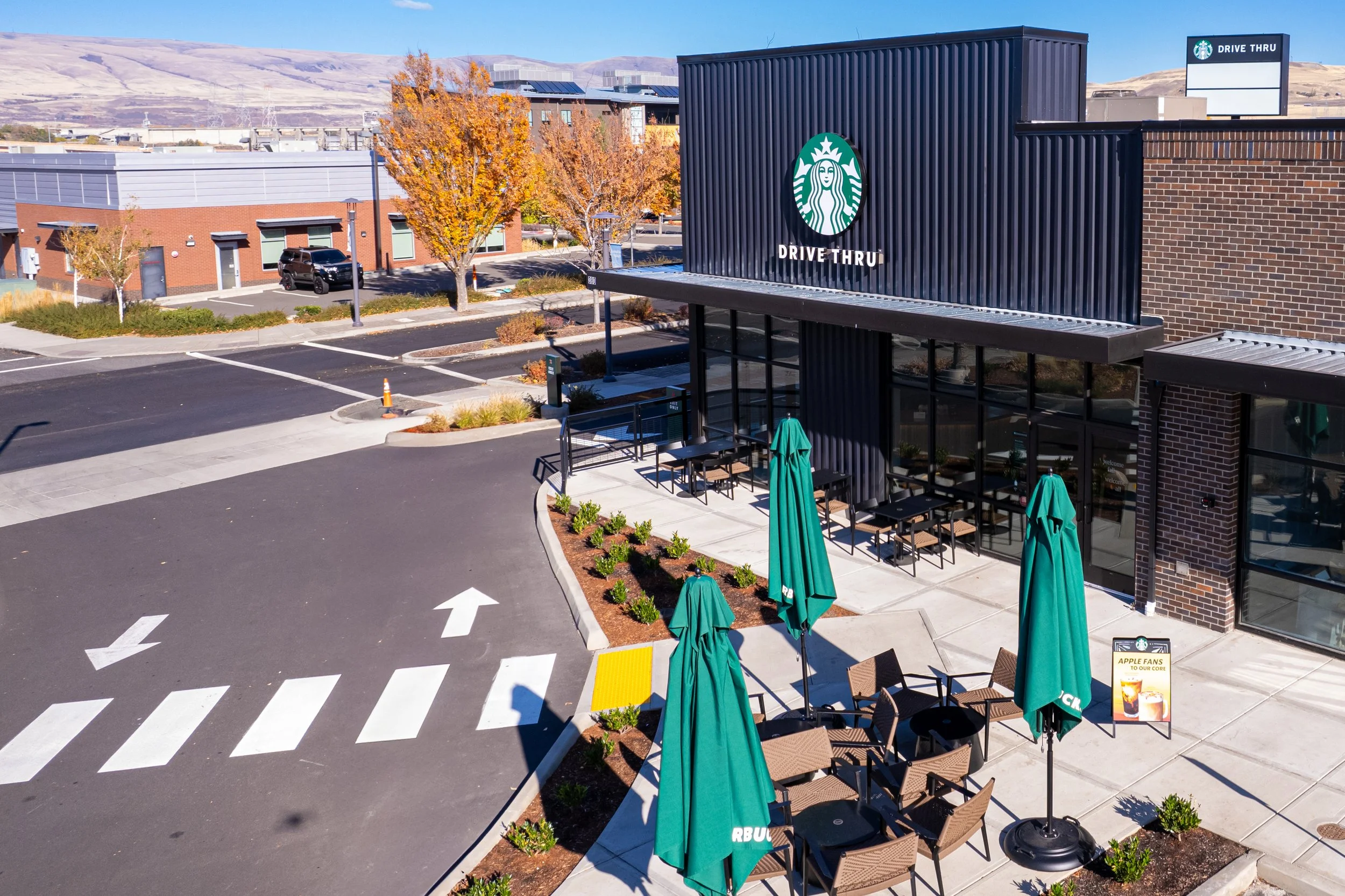 Exterior view of Starbucks drive-thru with outdoor seating area, green umbrellas, and a small sign promoting apple fans. Parking lot and nearby buildings are visible in the background.