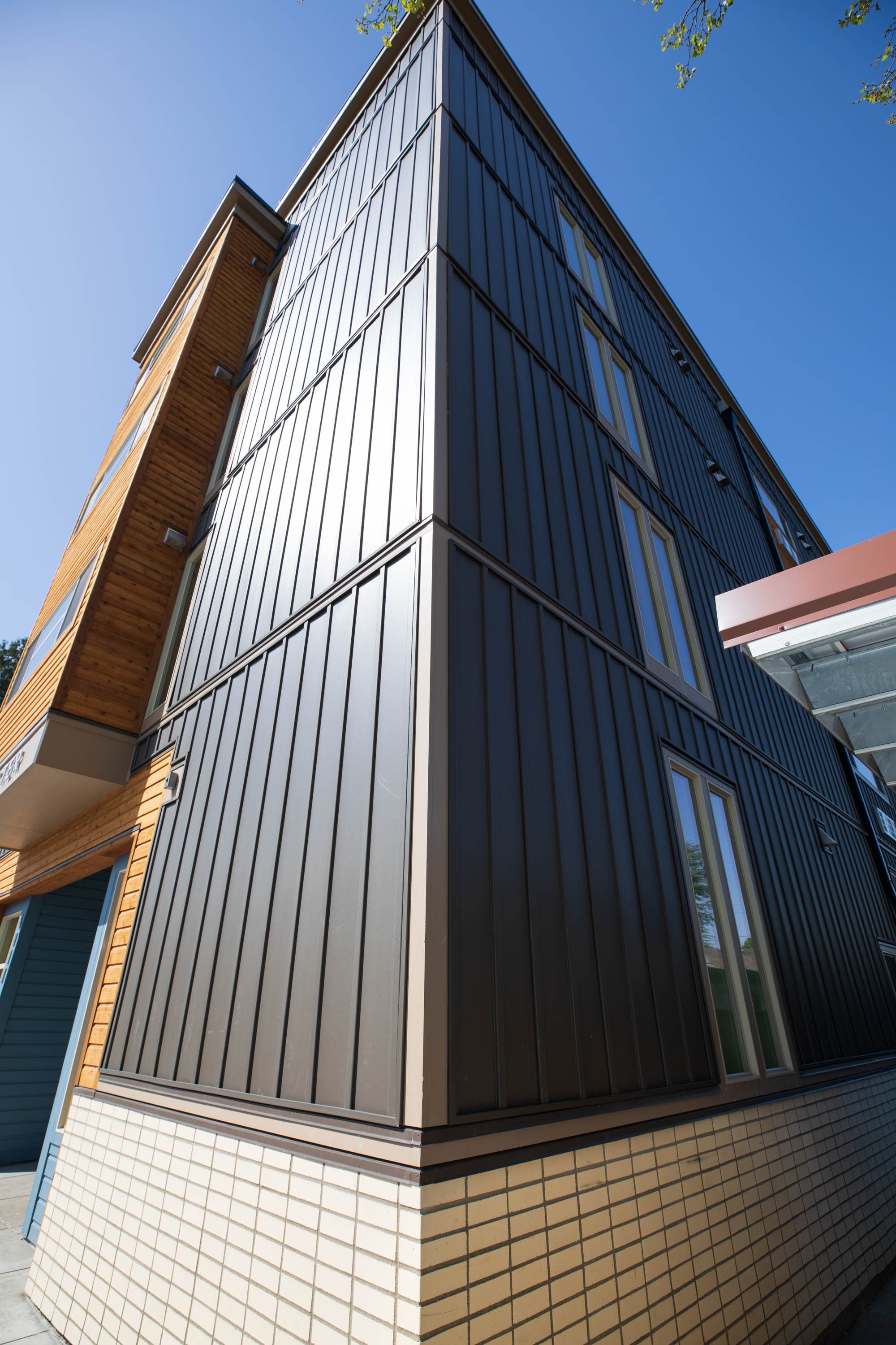 Low-angle view of a modern multi-story building with black metal siding, beige brick base, and small windows under a clear blue sky.