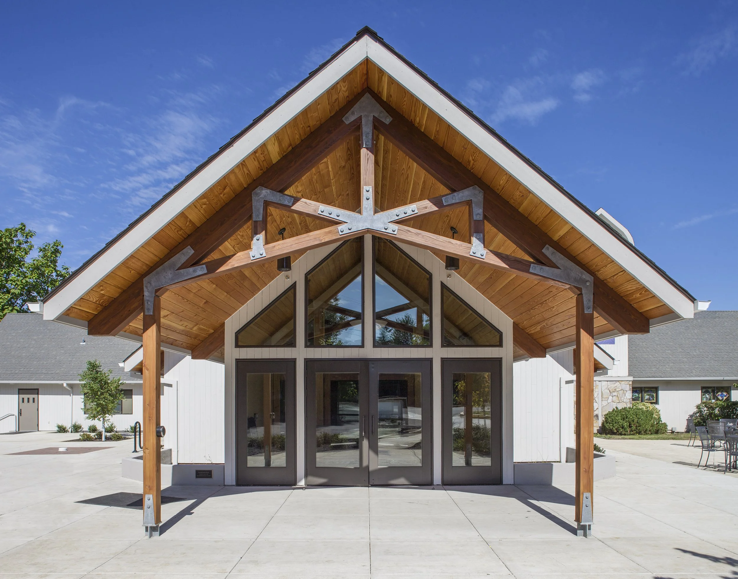 Front view of a modern building with large glass doors and a prominent wooden and metal A-frame roof under a clear blue sky.