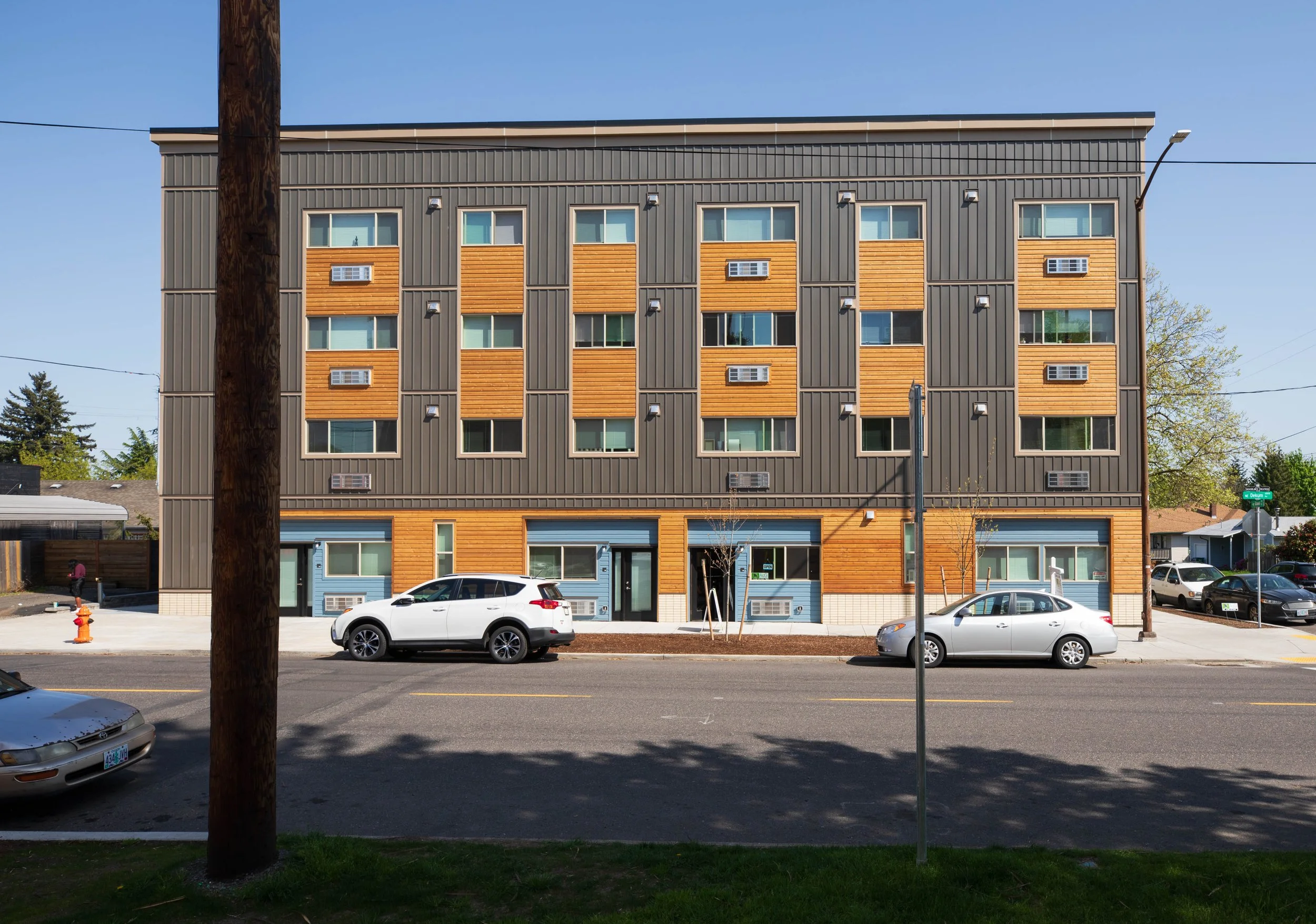 Modern multi-story residential building with brown and wooden exterior, multiple windows, air conditioning units, and parked cars in front.