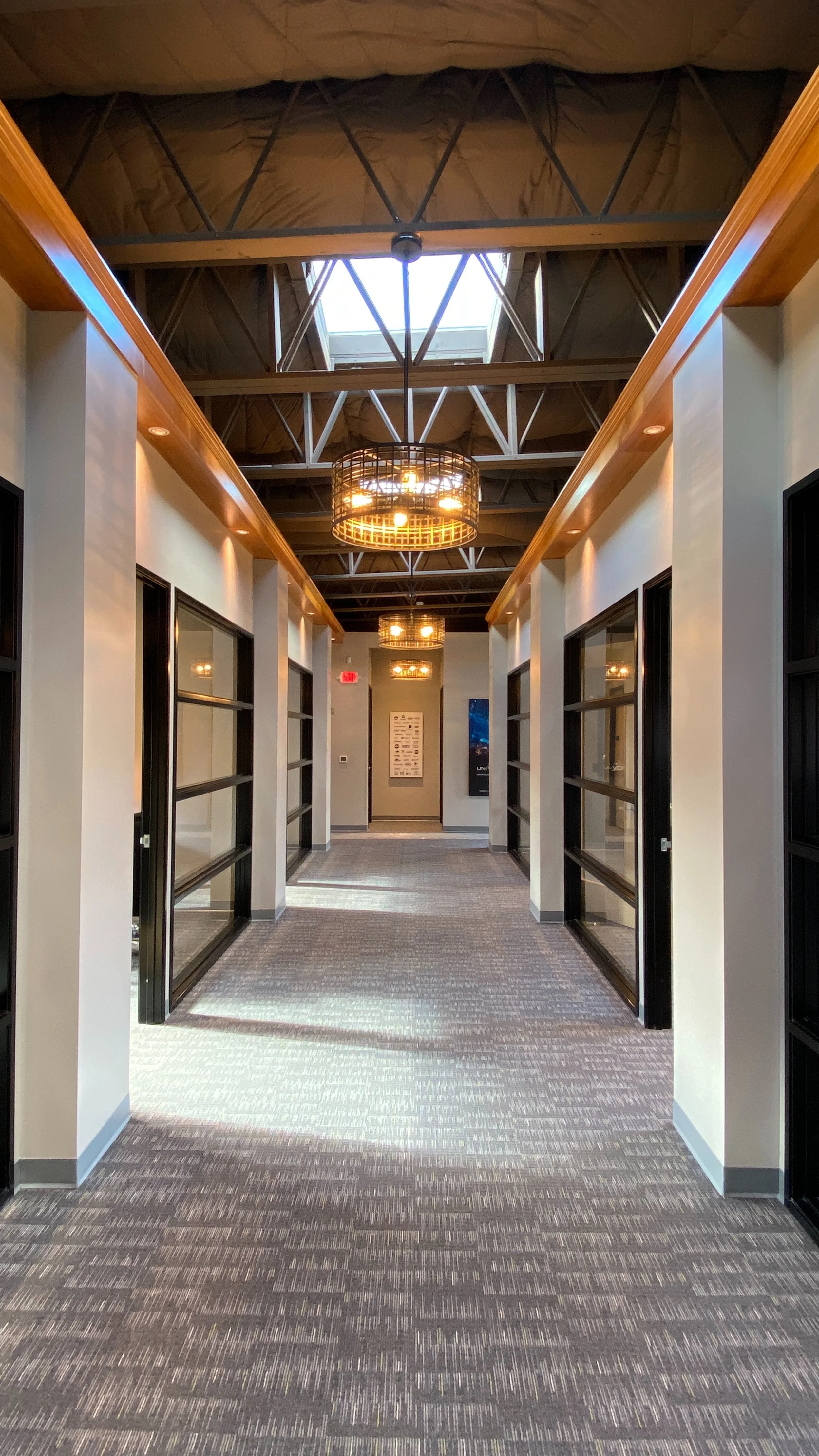 Empty corridor in modern office building with large windows, carpeted floor, and hanging circular light fixtures.