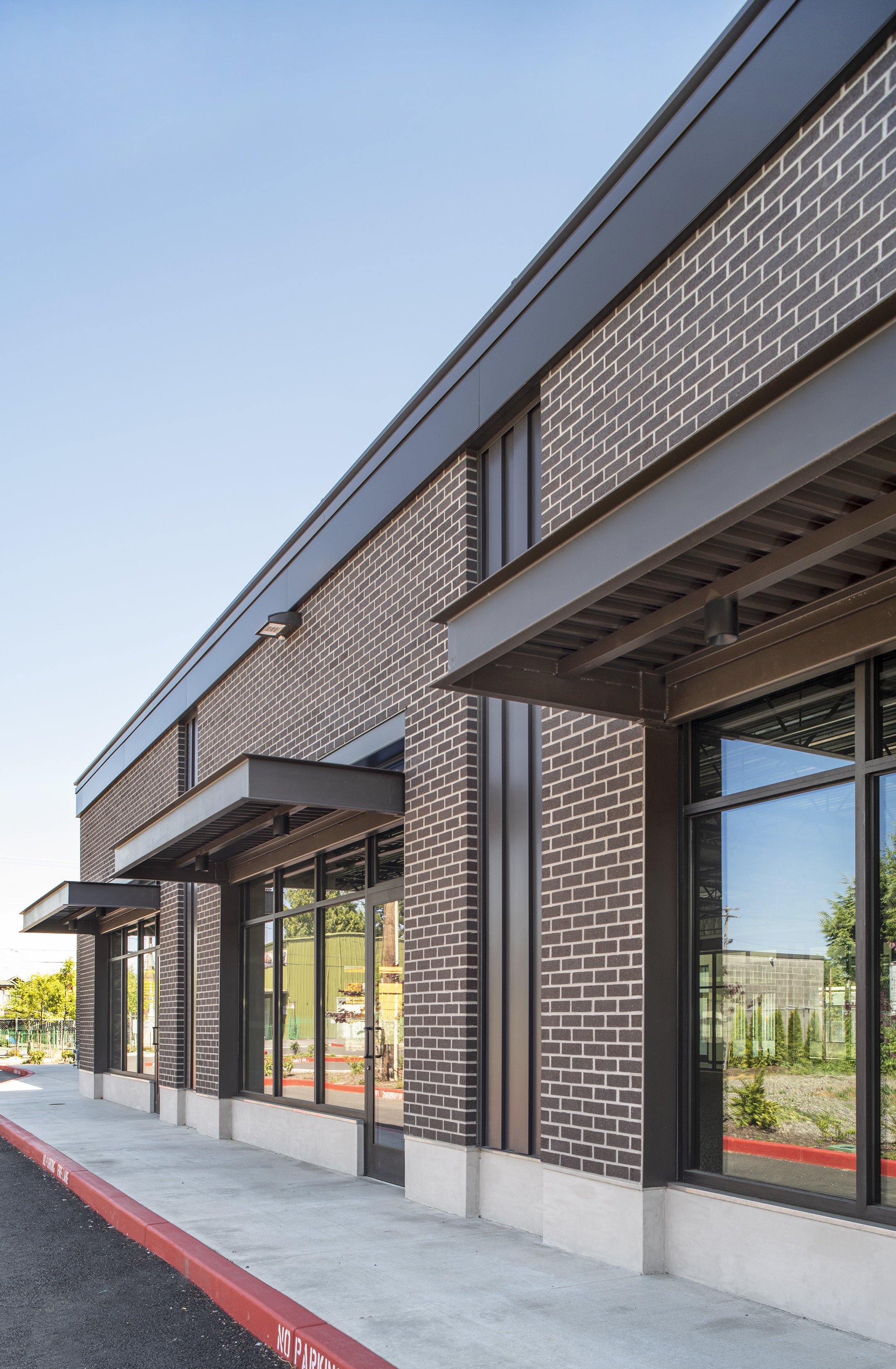 Modern commercial building with brick facade, large glass windows, and small overhangs, near a sidewalk and parking lot.