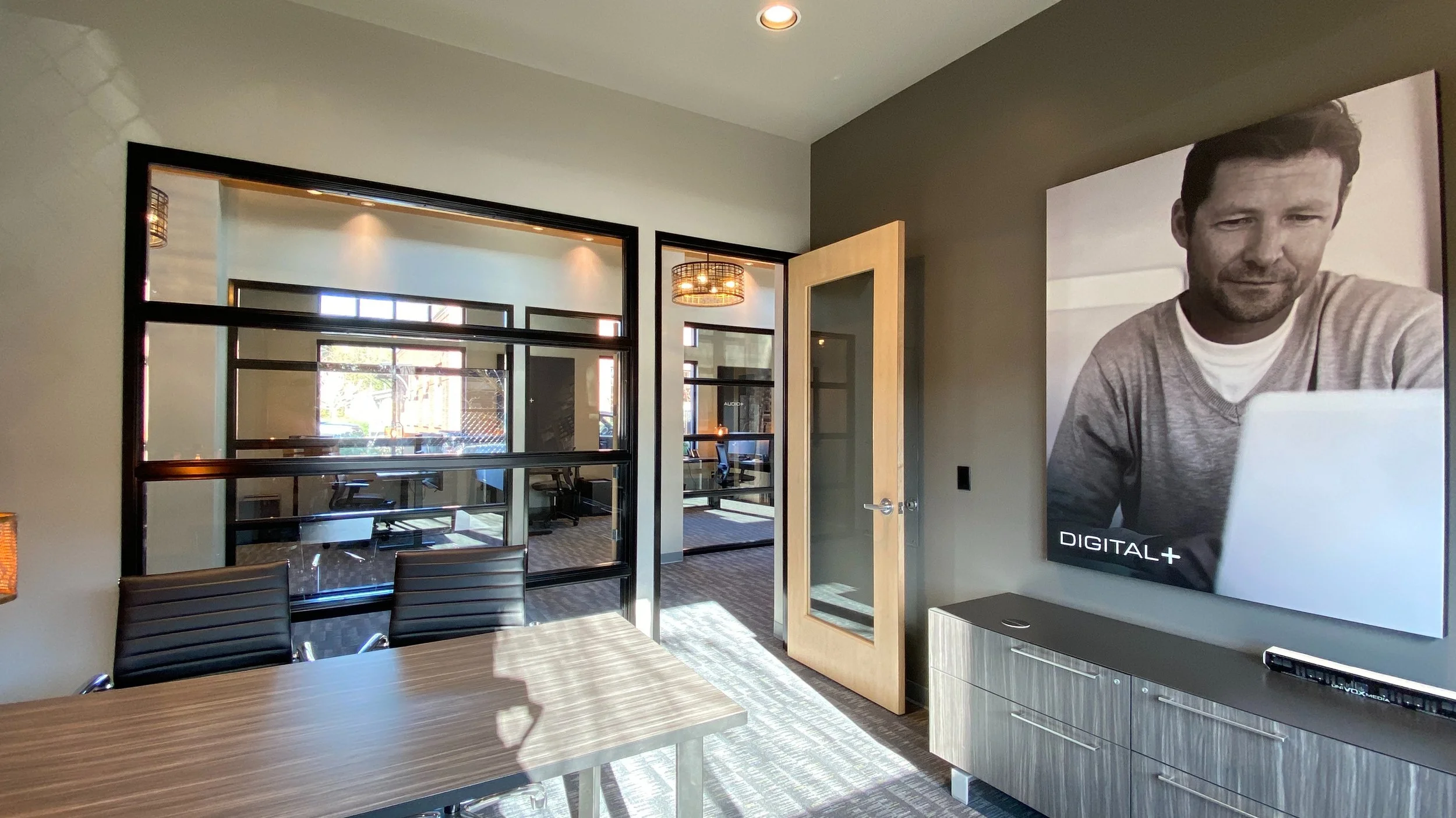 Indoor office conference room with a wooden table, black chairs, large windows, a glass door, and a wall-mounted TV showing a black-and-white photo of a man using a laptop.