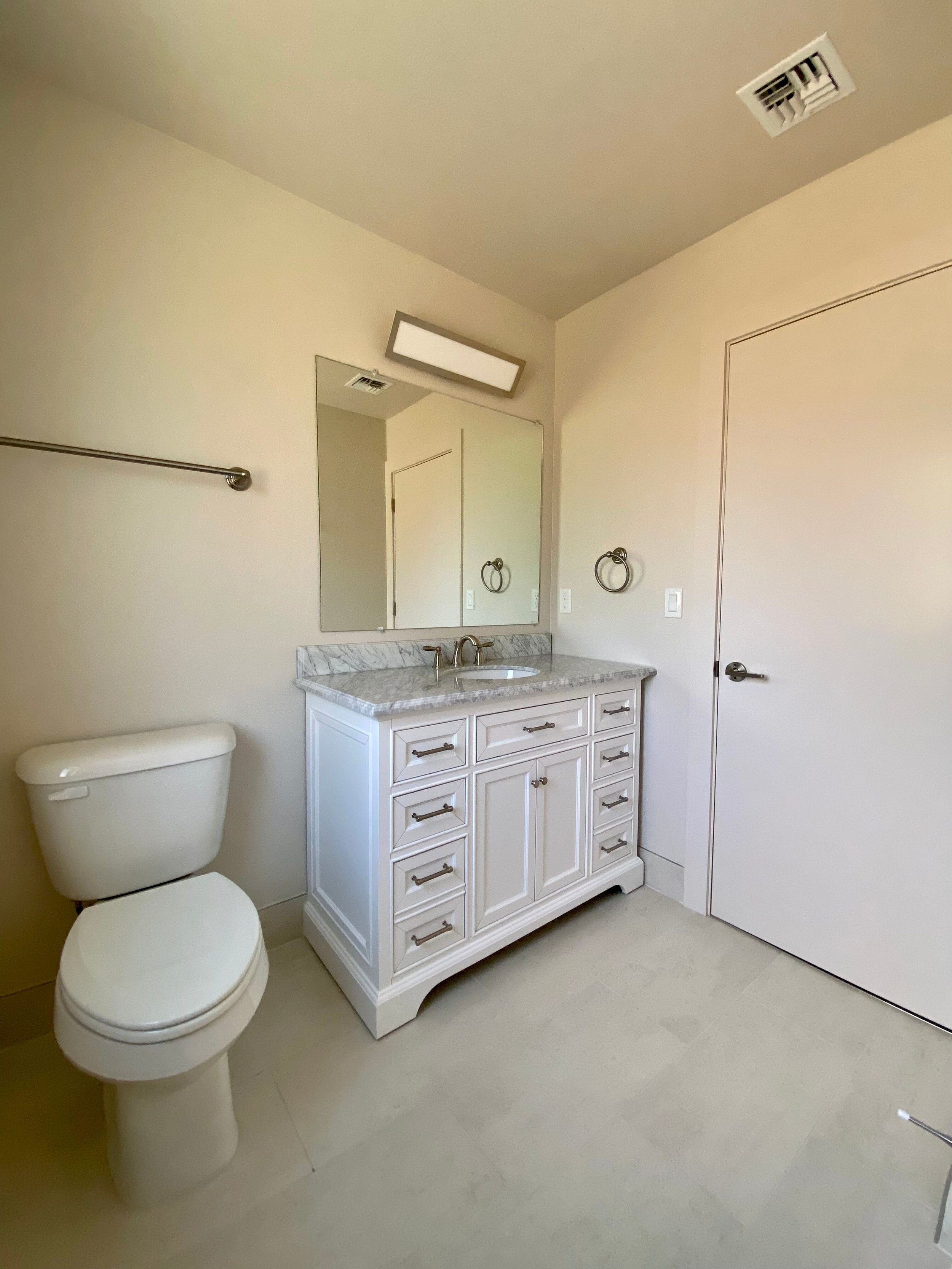 A bathroom with a white toilet, a white vanity with a marble countertop, a mirror above the vanity, and beige walls.