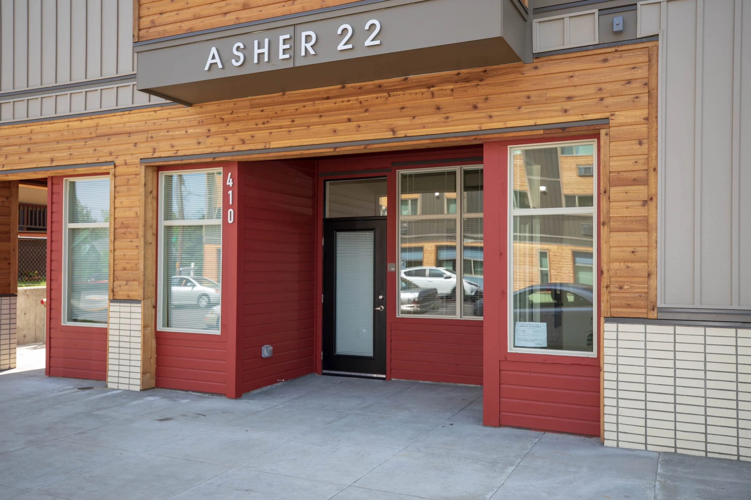 Exterior view of a modern building with wood and gray siding, red accents around the door, and large windows. The sign reads 'ASHE 22' above the entrance, and the street number '410' is on the wall next to the door.