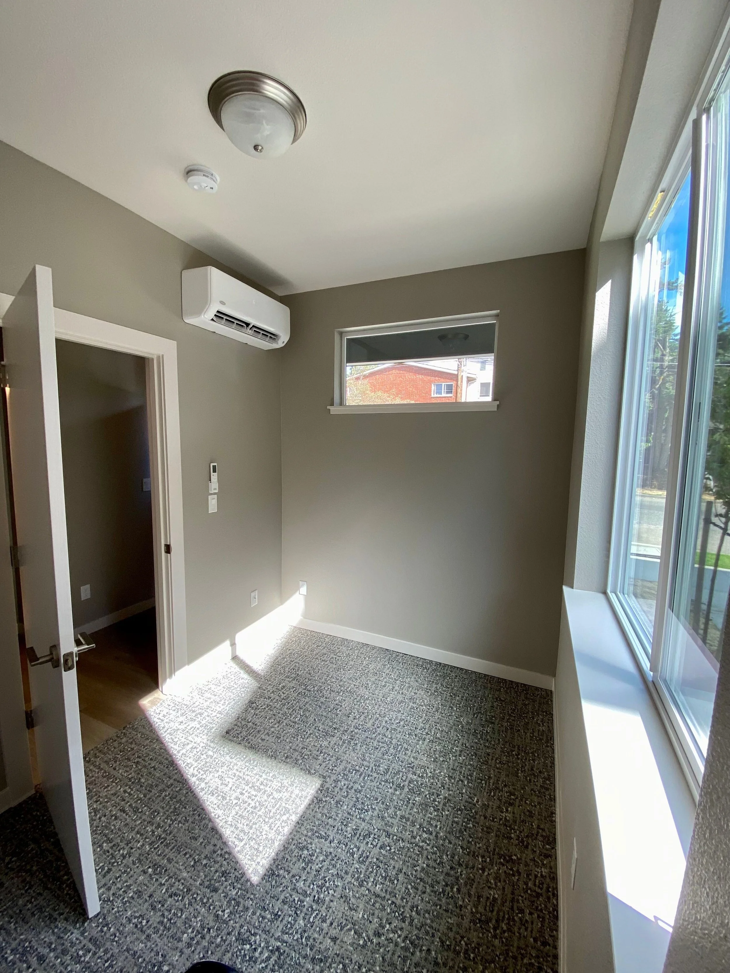 Empty small room with gray walls, a window, carpeted floor, ceiling light, and wall-mounted heater.