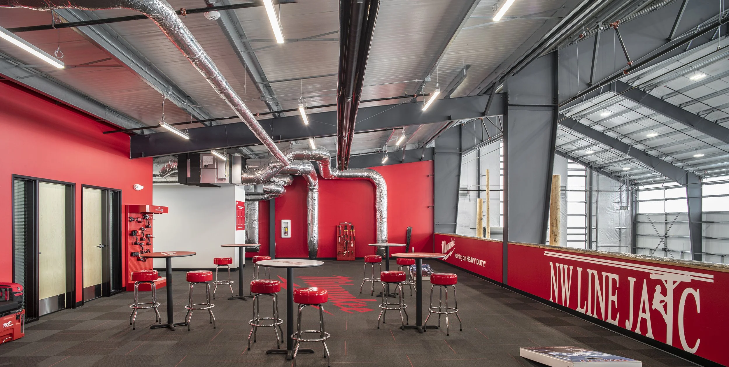 Indoor sports facility or training area with red and gray walls, high ceiling, exposed ducts and pipes, and multiple round tables with red stools, featuring branding for Milwaukee and NW Line JAC.