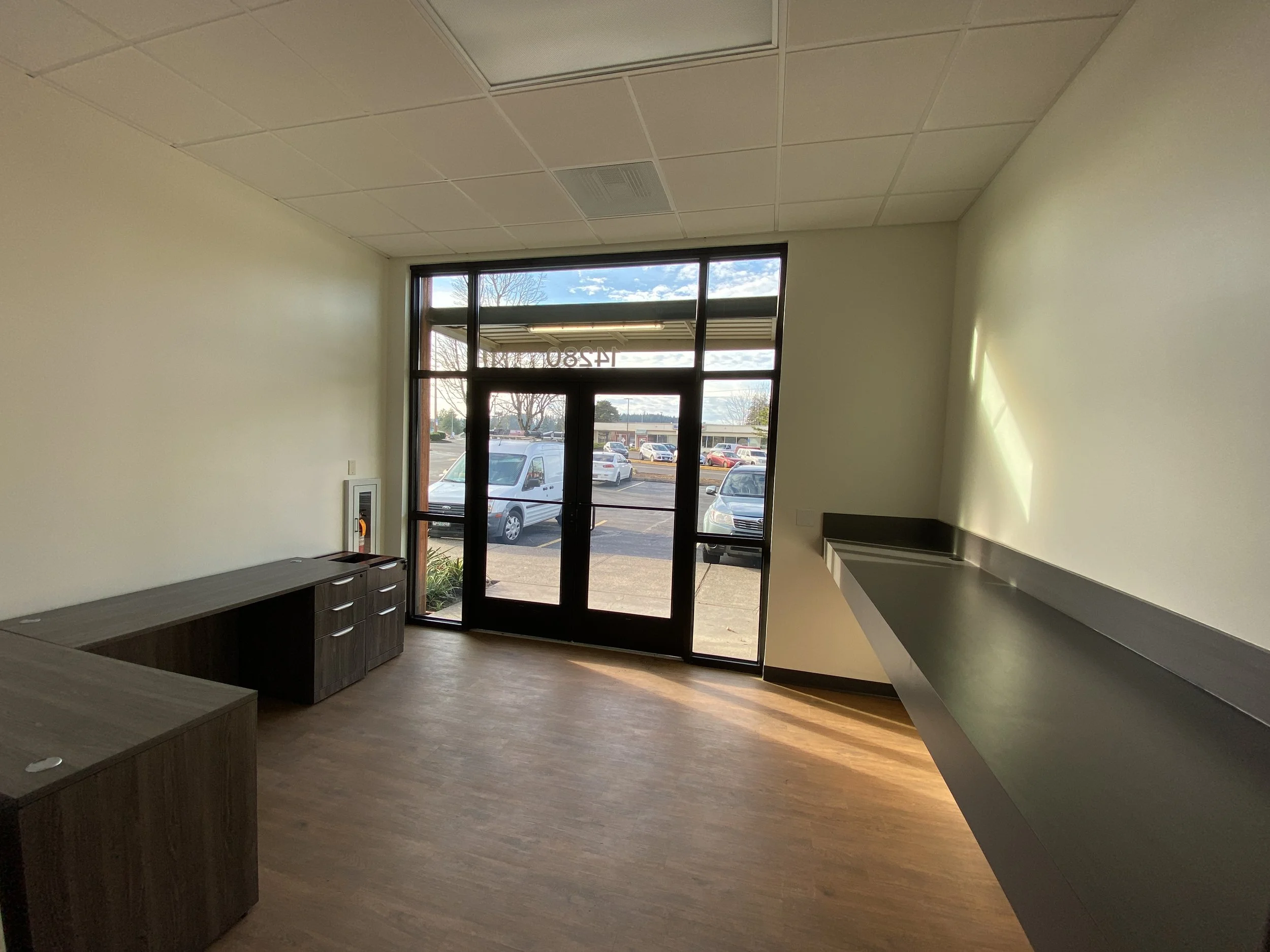An empty room with a large glass front door and windows, a wooden floor, a dark-colored countertop on the right wall, and a wooden desk on the left side near the wall. Outside the window, there are parked cars and trees in a parking lot.
