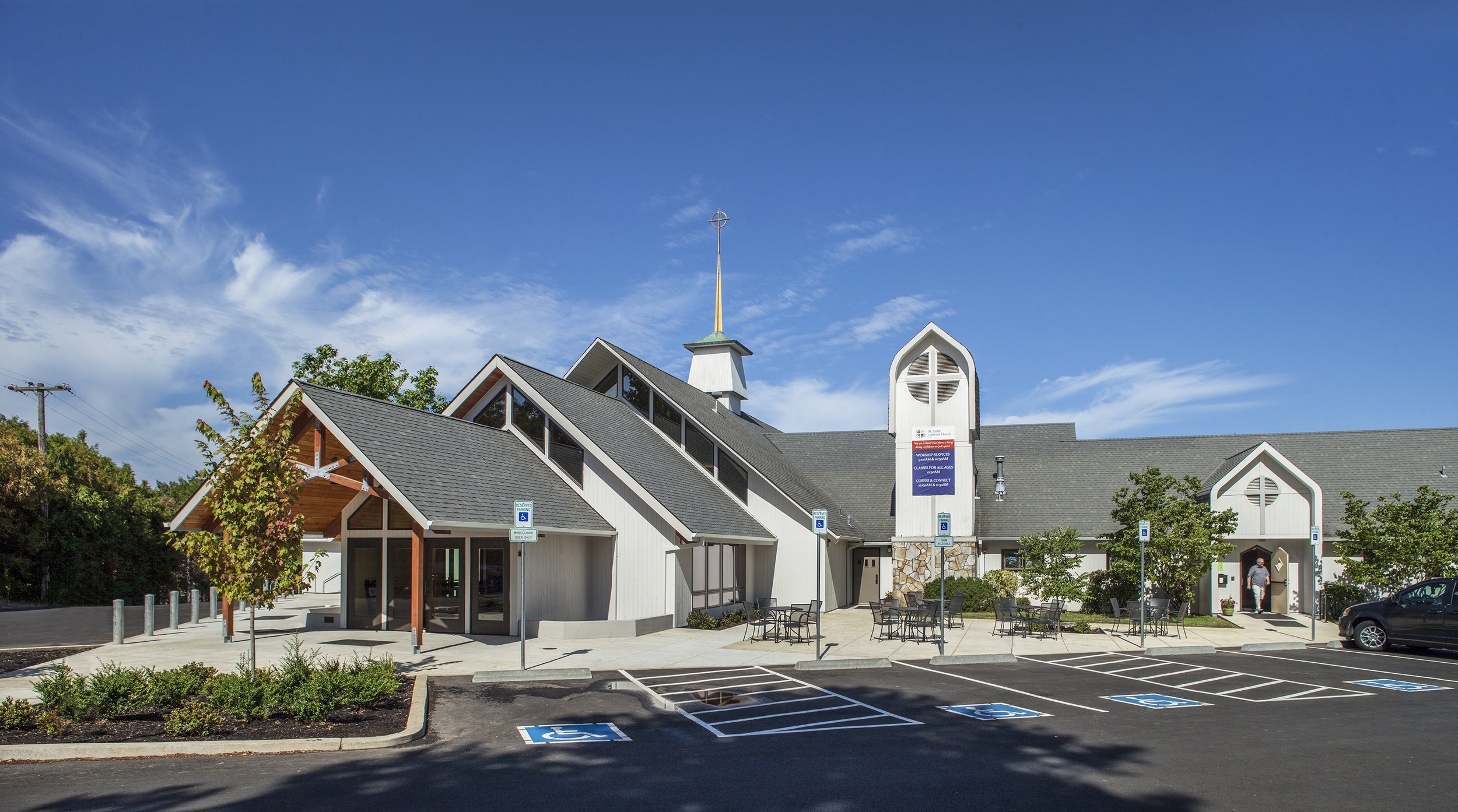 A modern church building with a parking lot in front, including designated accessible parking spaces, surrounded by trees and sitting under a blue sky with some clouds.