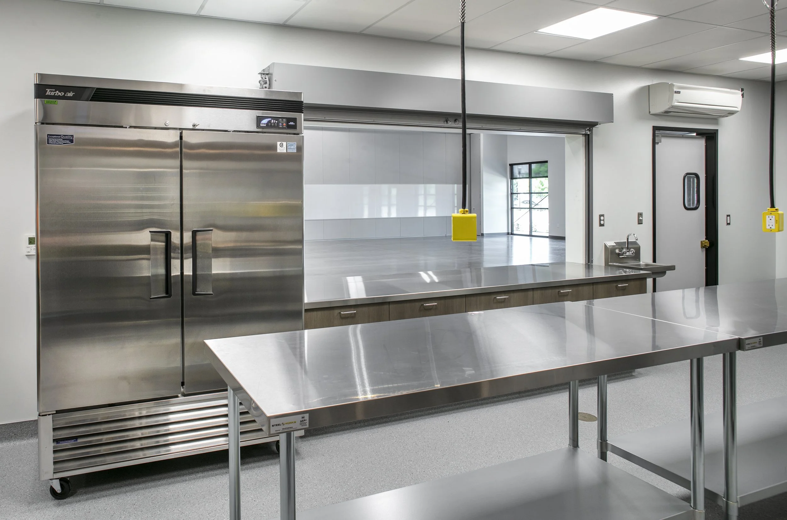 Commercial kitchen with stainless steel refrigerator, stainless steel tables, and a serving area window.