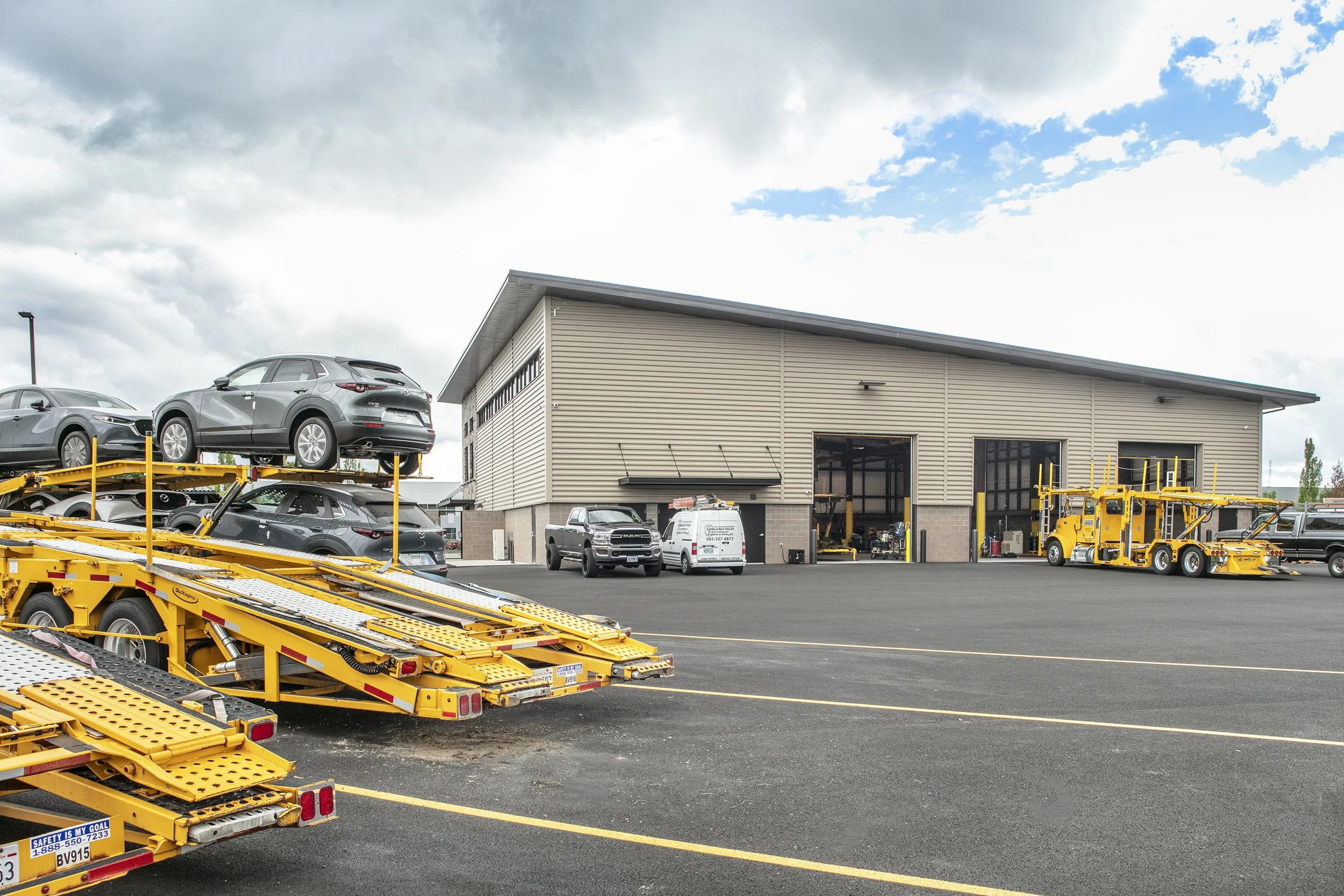 Car dealership or service center with vehicles on yellow car transport trailers and parked on black asphalt lot in front of large industrial building with open garage bays, cloudy sky overhead.