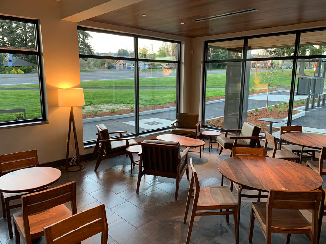 A cozy cafe or lounge with wooden tables and chairs, large windows showing a view of a grassy outdoor area and a sidewalk outside, a floor lamp providing warm light.