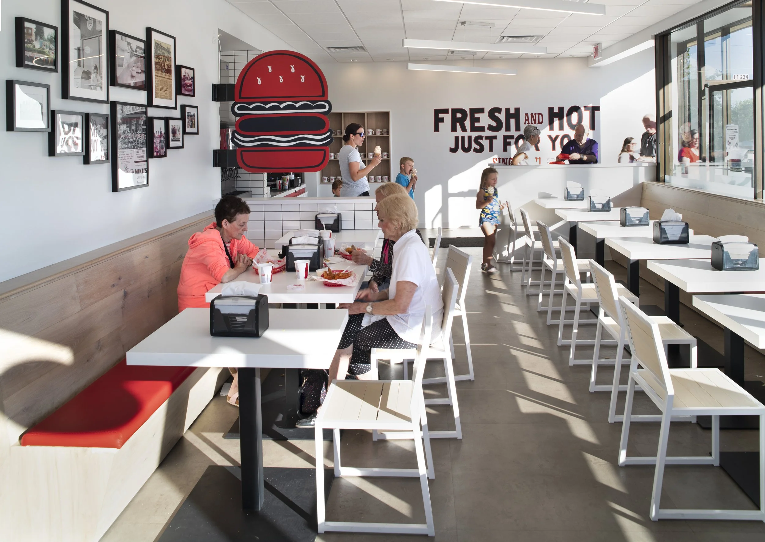 Inside a fast food restaurant with white tables and chairs, some customers eating, others waiting in line, large 'FRESH AND HOT JUST FOR YOU' sign on the wall, a wall art display with framed pictures, and a large burger graphic hanging from the ceili