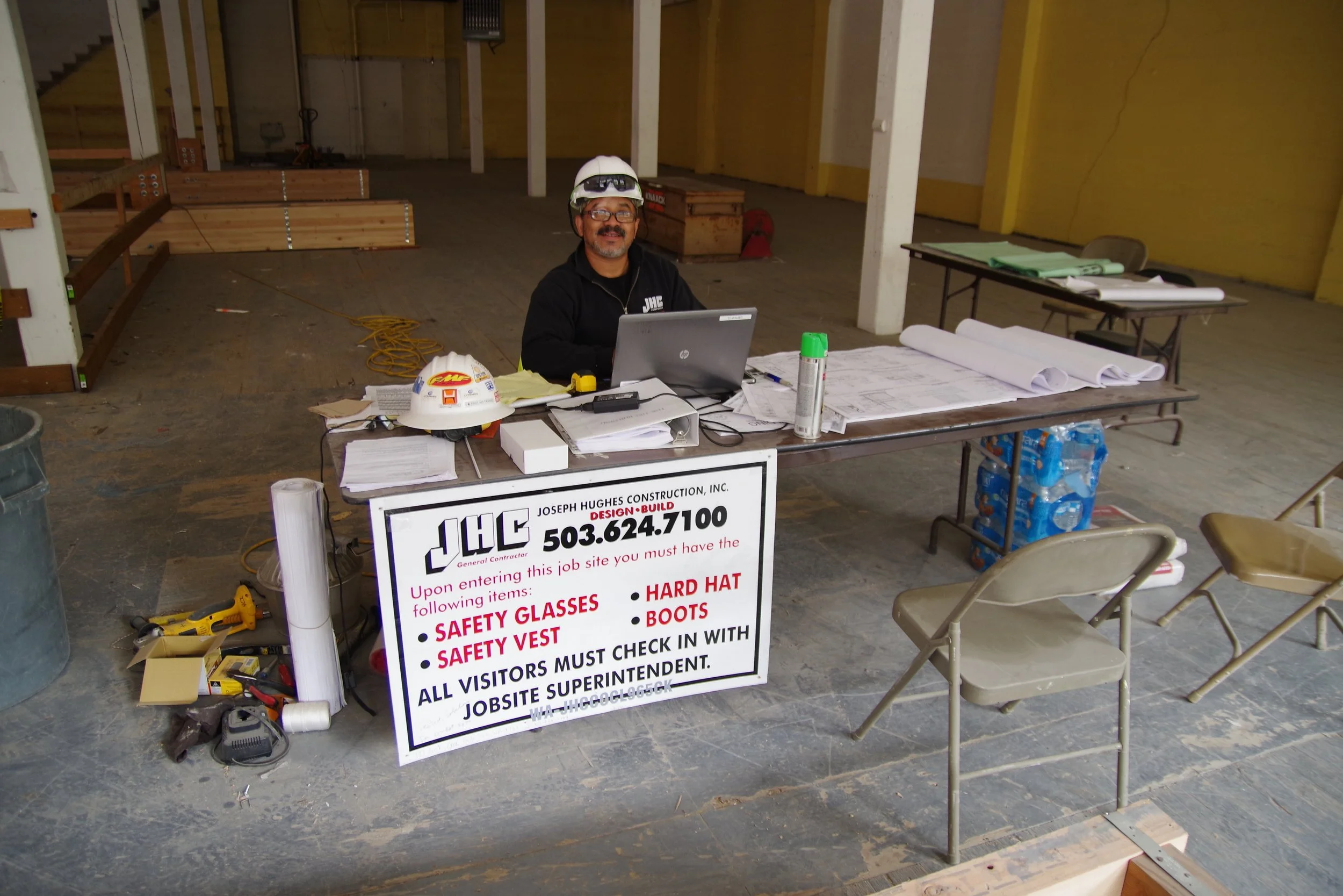 Construction site supervisor sitting at a table with safety gear, plans, and a laptop, inside a partially built building.