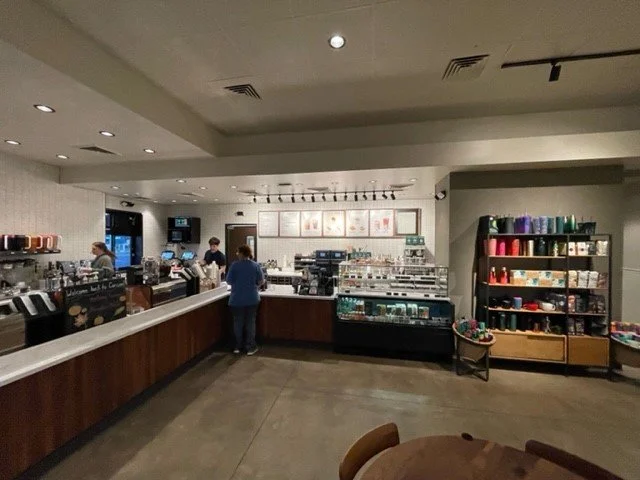 Interior of a coffee shop with baristas preparing drinks, a display case with baked goods, and shelves with colorful mugs and merchandise.