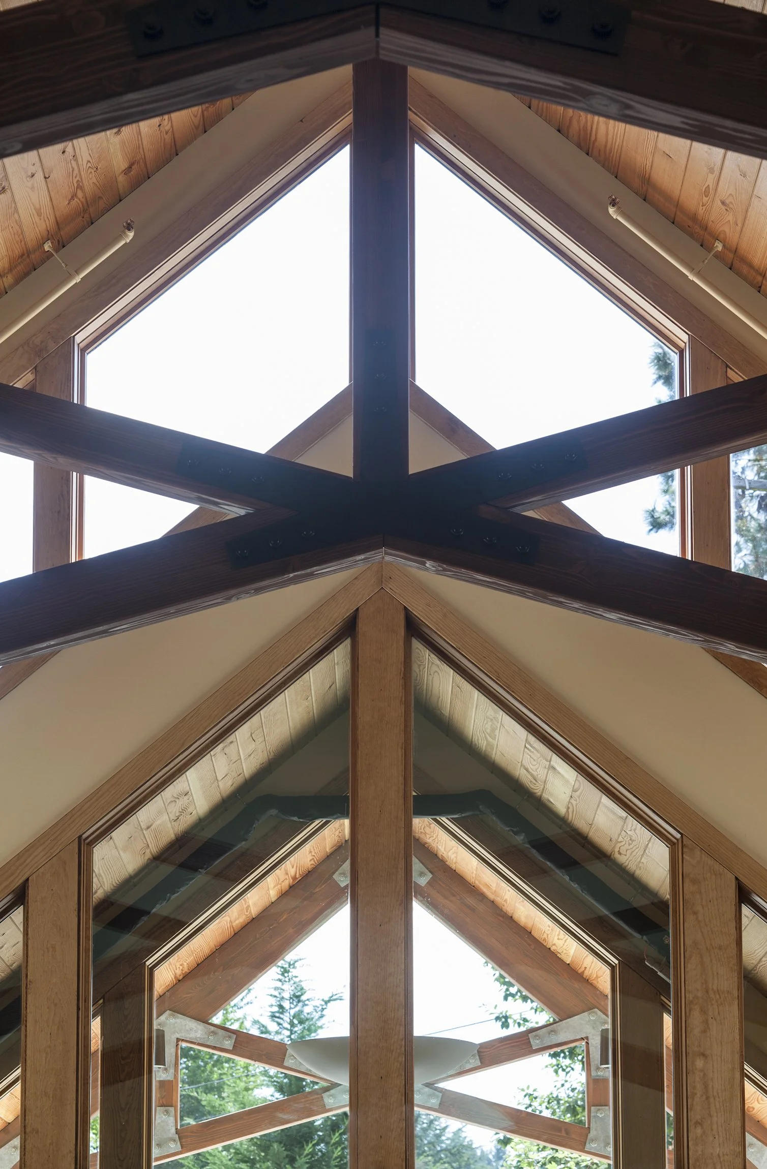 View of a wooden A-frame roof with large glass windows and beams, showing an outdoor scene with trees and a cloudy sky through the windows.