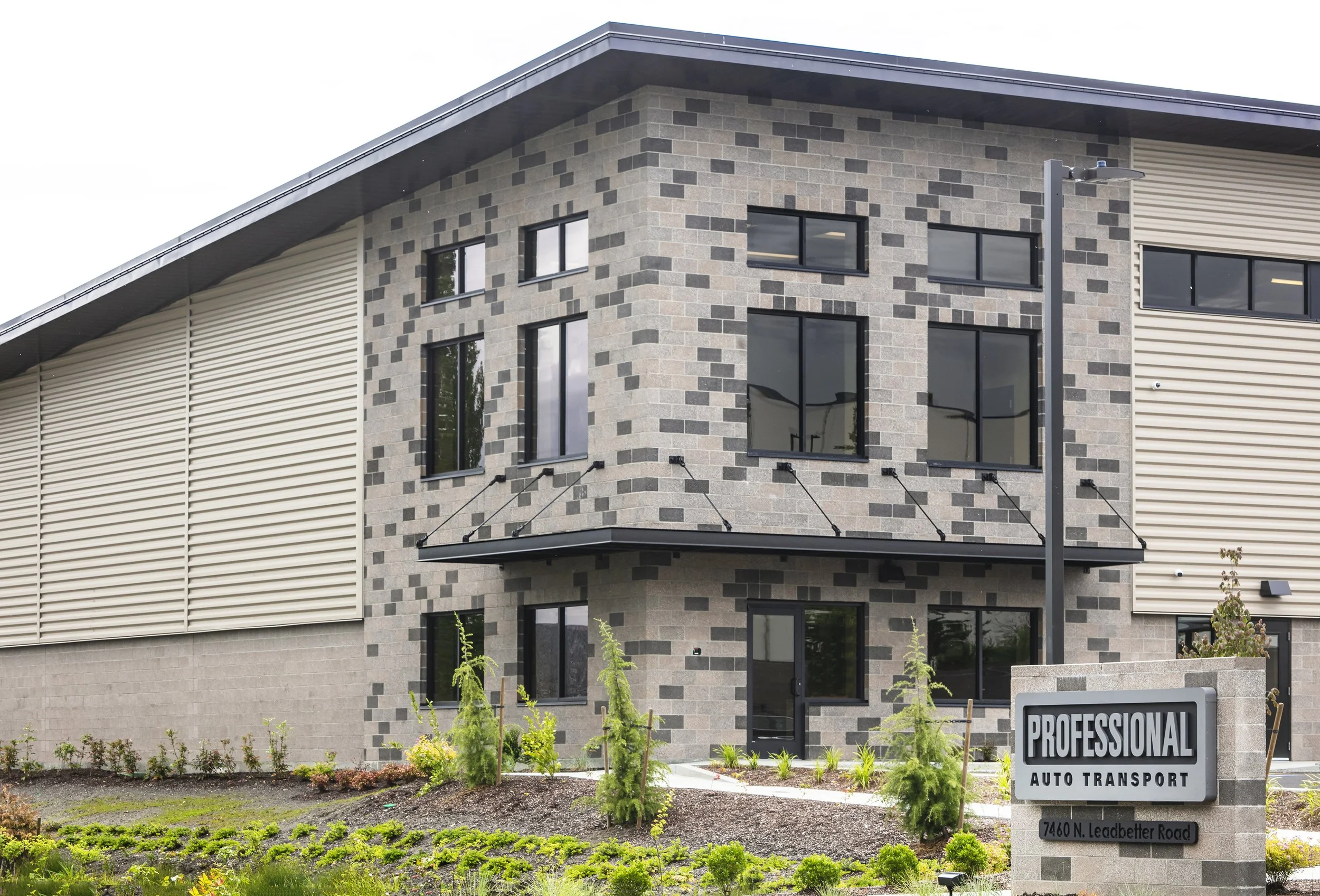 Modern two-story commercial building with a brick facade, large windows, and a landscaped front yard featuring small trees and bushes. A sign reads 'Professional Auto Transport' with the address '7460 N. Leadbetter Road'.