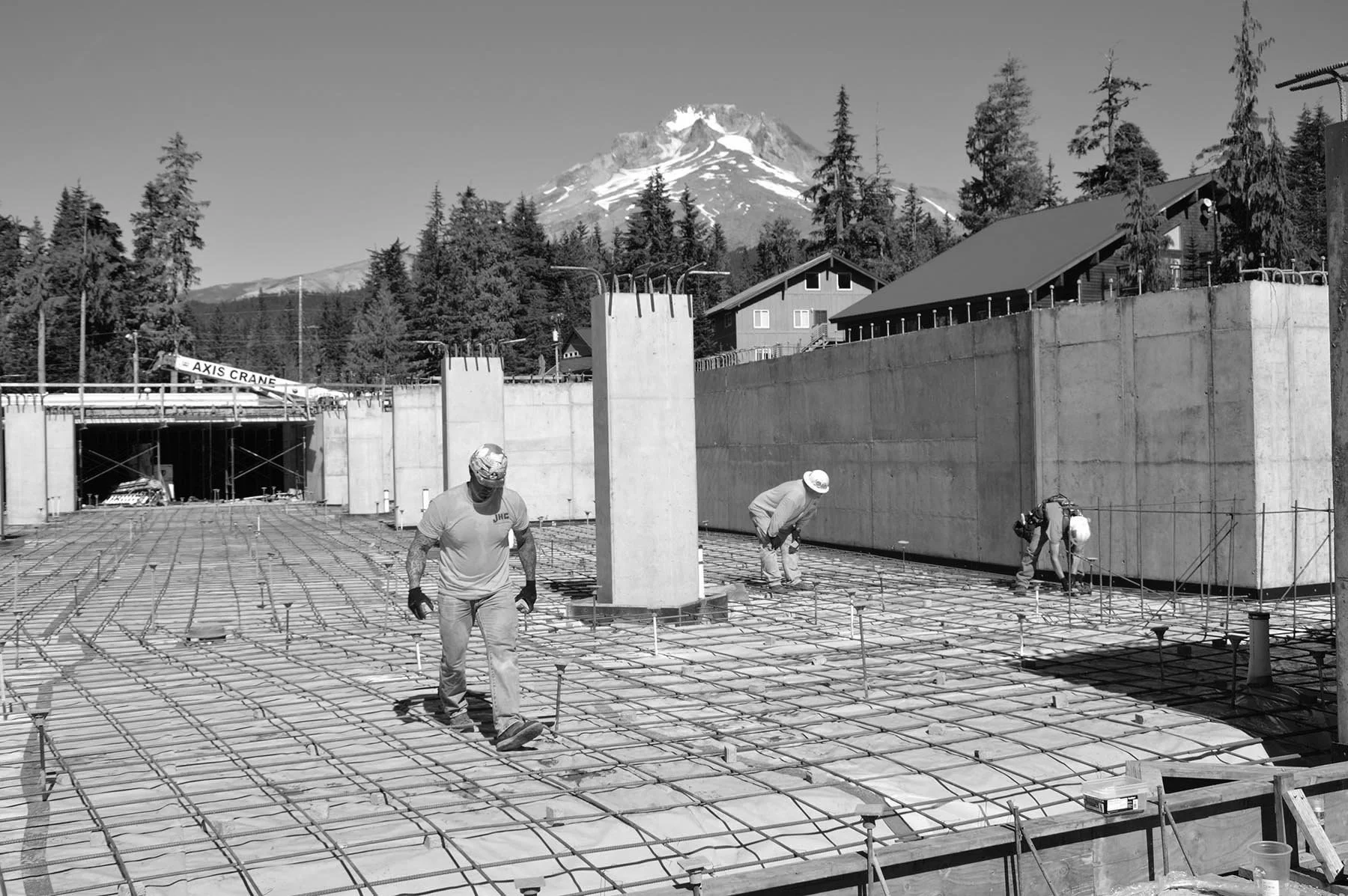 Construction workers are working on a concrete building foundation with rebar, with a mountain and trees in the background.