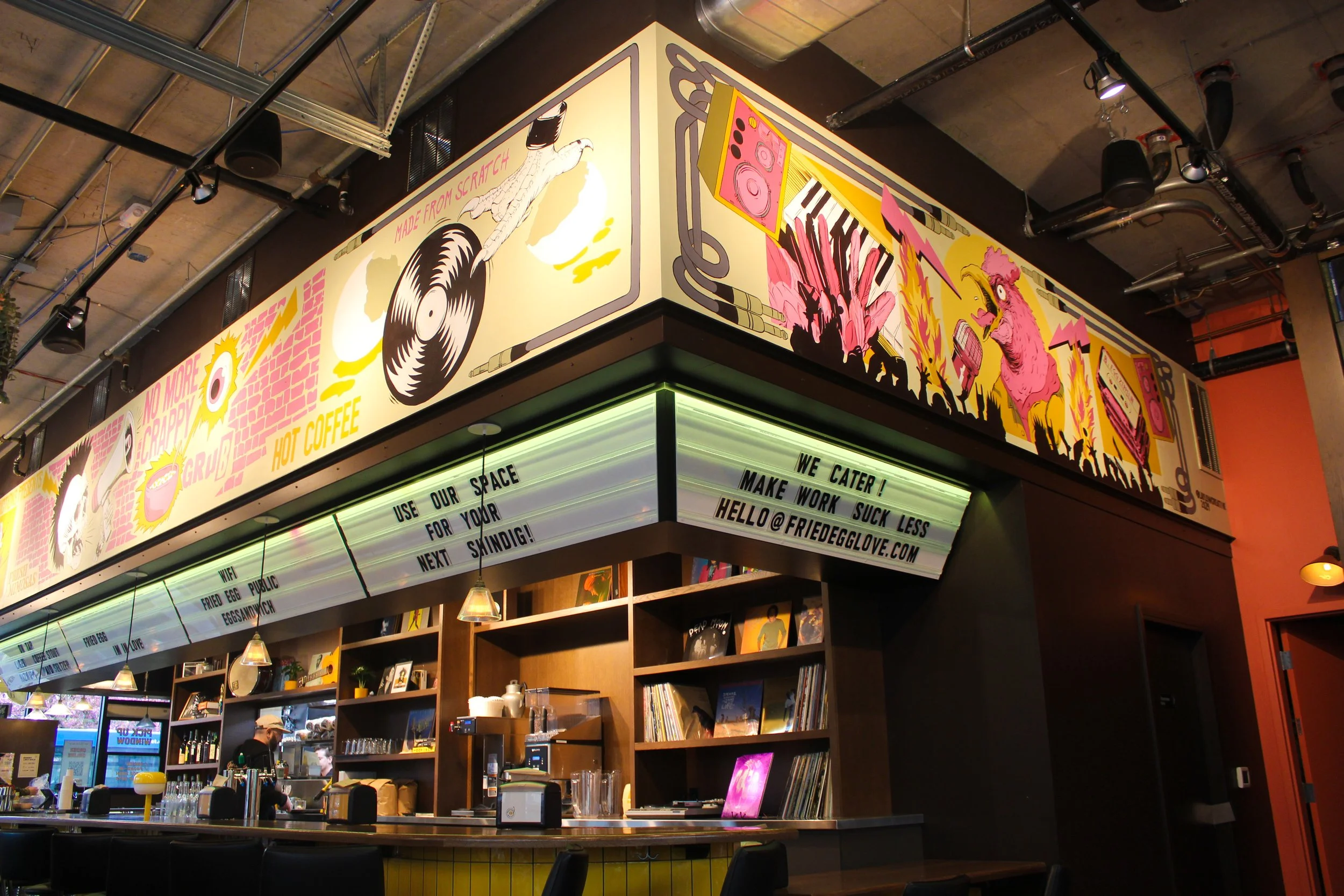 Colorful mural with images of a turntable, speaker, flamingos, and a pink dinosaur on the cafe wall, with a bar counter below and a menu board with messages about WiFi, fried eggs, and coffee.