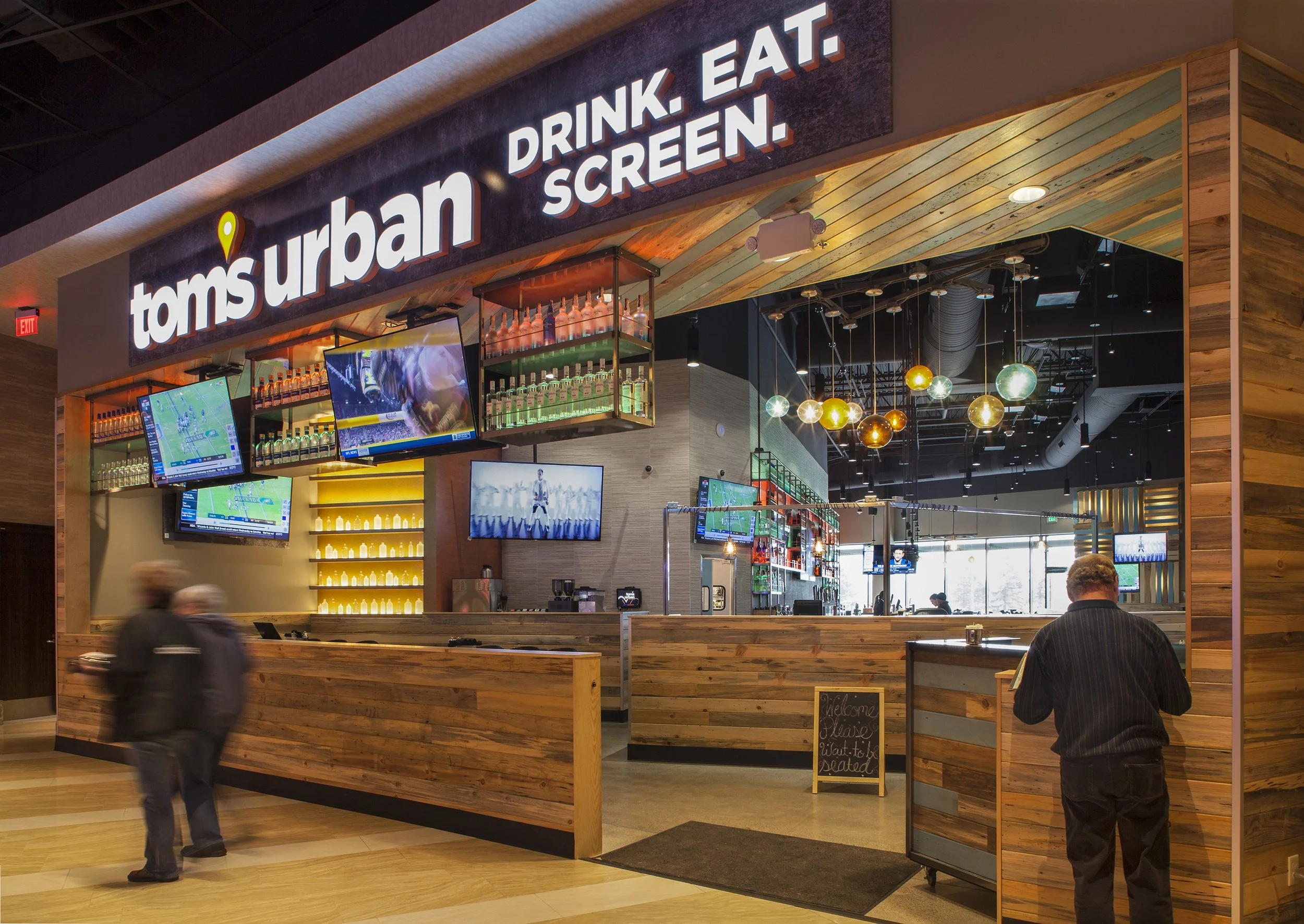 Tom's Urban restaurant with a wooden counter, multiple flat-screen TVs showing sports, neon sign reading 'Drink. Eat. Screen.', and colorful hanging lights inside a mall.