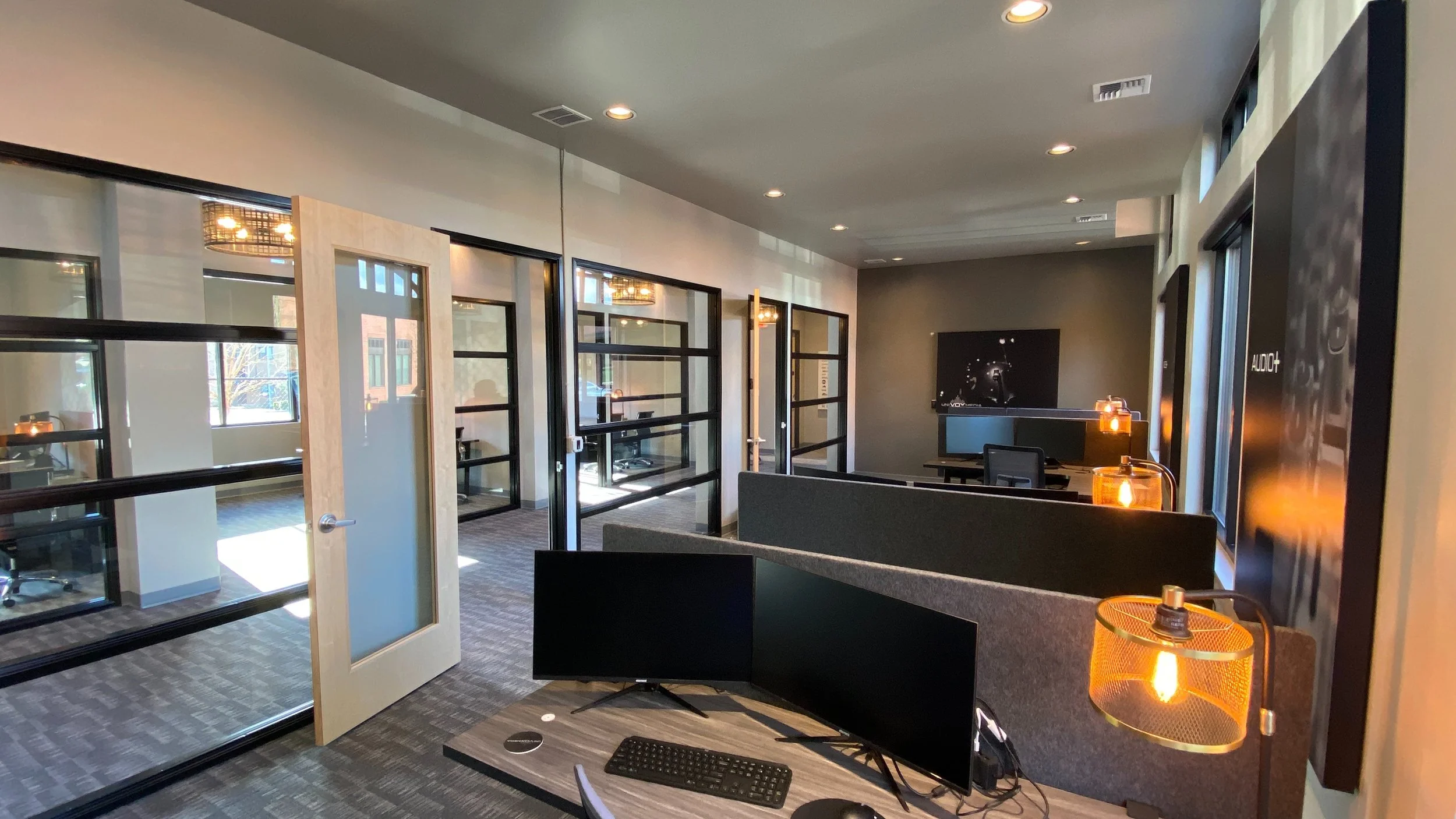 Modern office with glass-walled private rooms, some with desks and chairs, open workspace with dual computer monitors, cozy lighting fixtures, and a tv on the back wall.