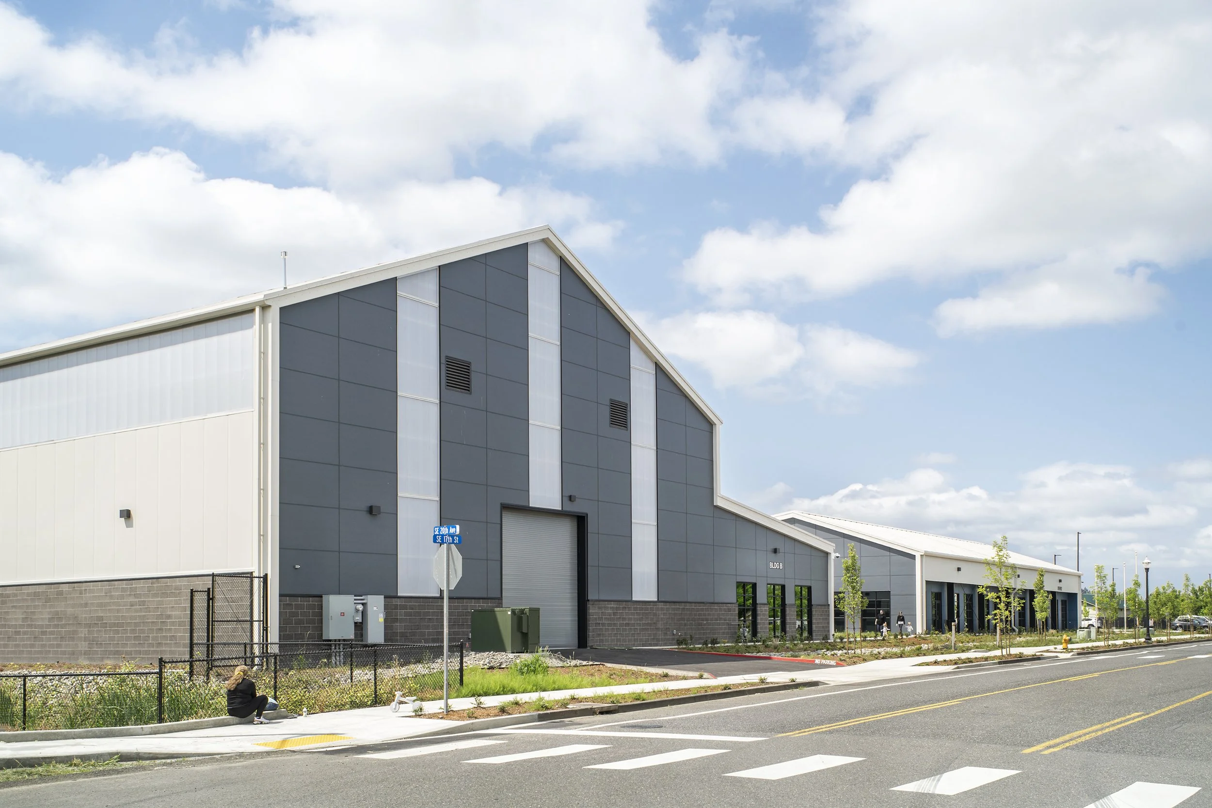Modern industrial building with a blue and white exterior, small trees lining the sidewalk, and a woman sitting on the curb on a sunny day.
