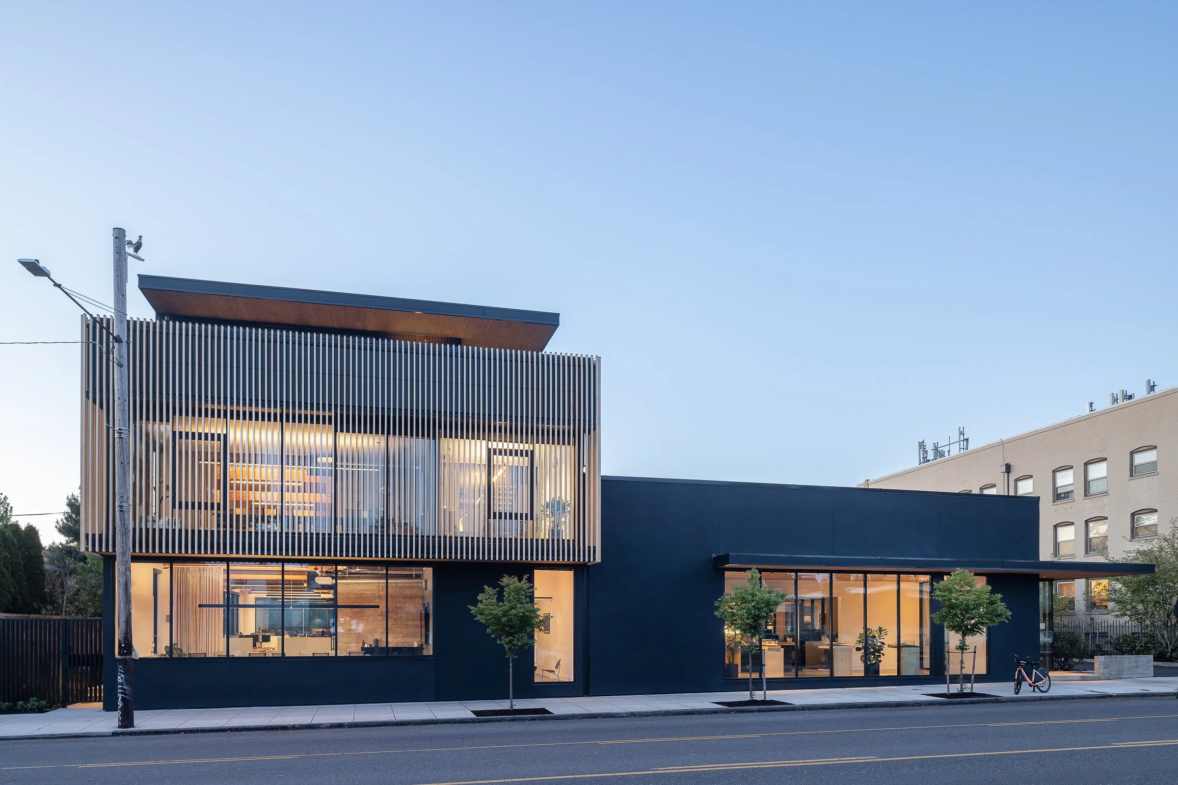 Modern two-story building with large glass windows, blue exterior, and vertical wooden slats on the upper level. Small trees line the sidewalk in front. The photo is taken during twilight.