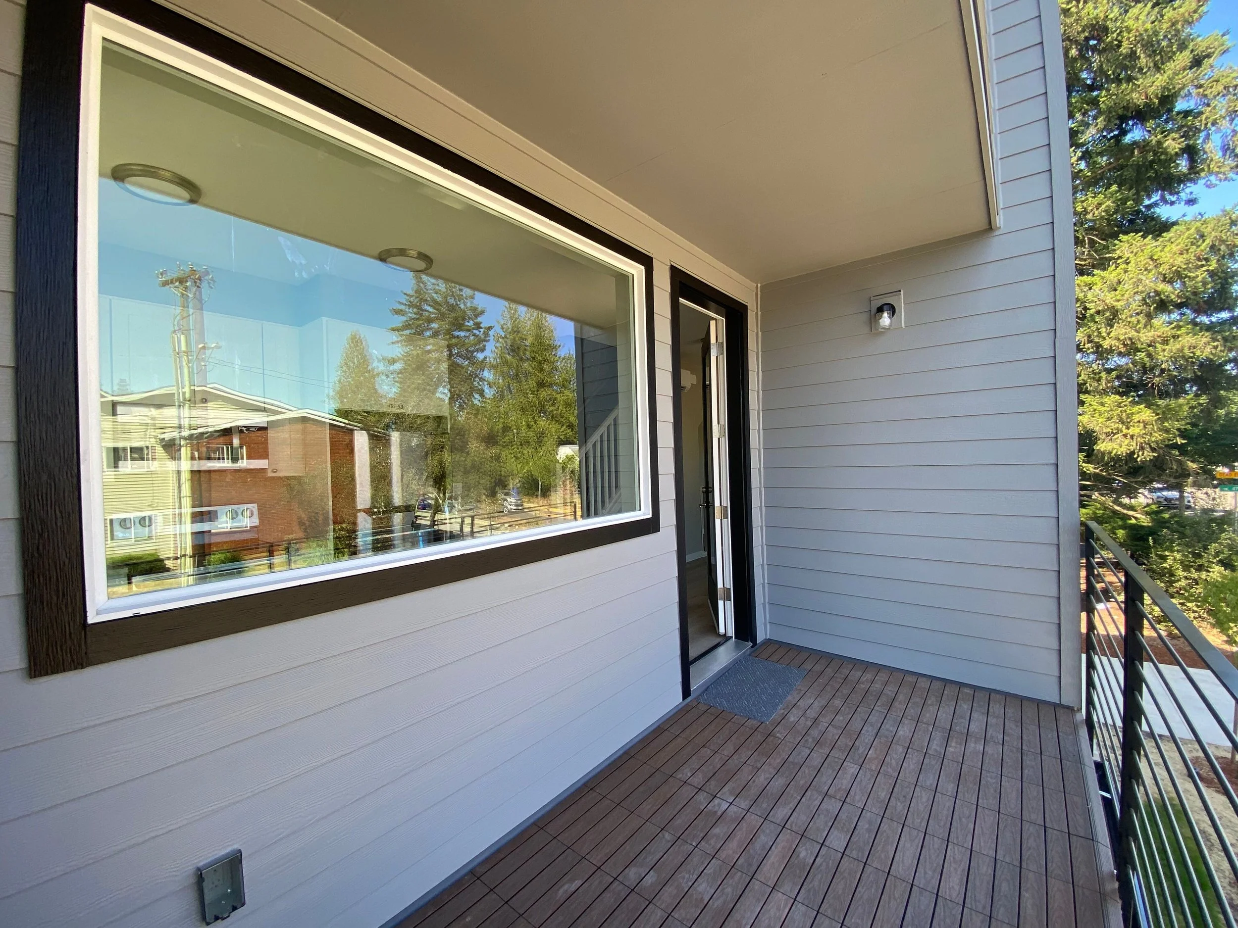 View of a modern apartment balcony with wooden flooring, a large window, a glass door, and an outdoor wall light fixture. Trees and neighboring buildings are reflected in the window.