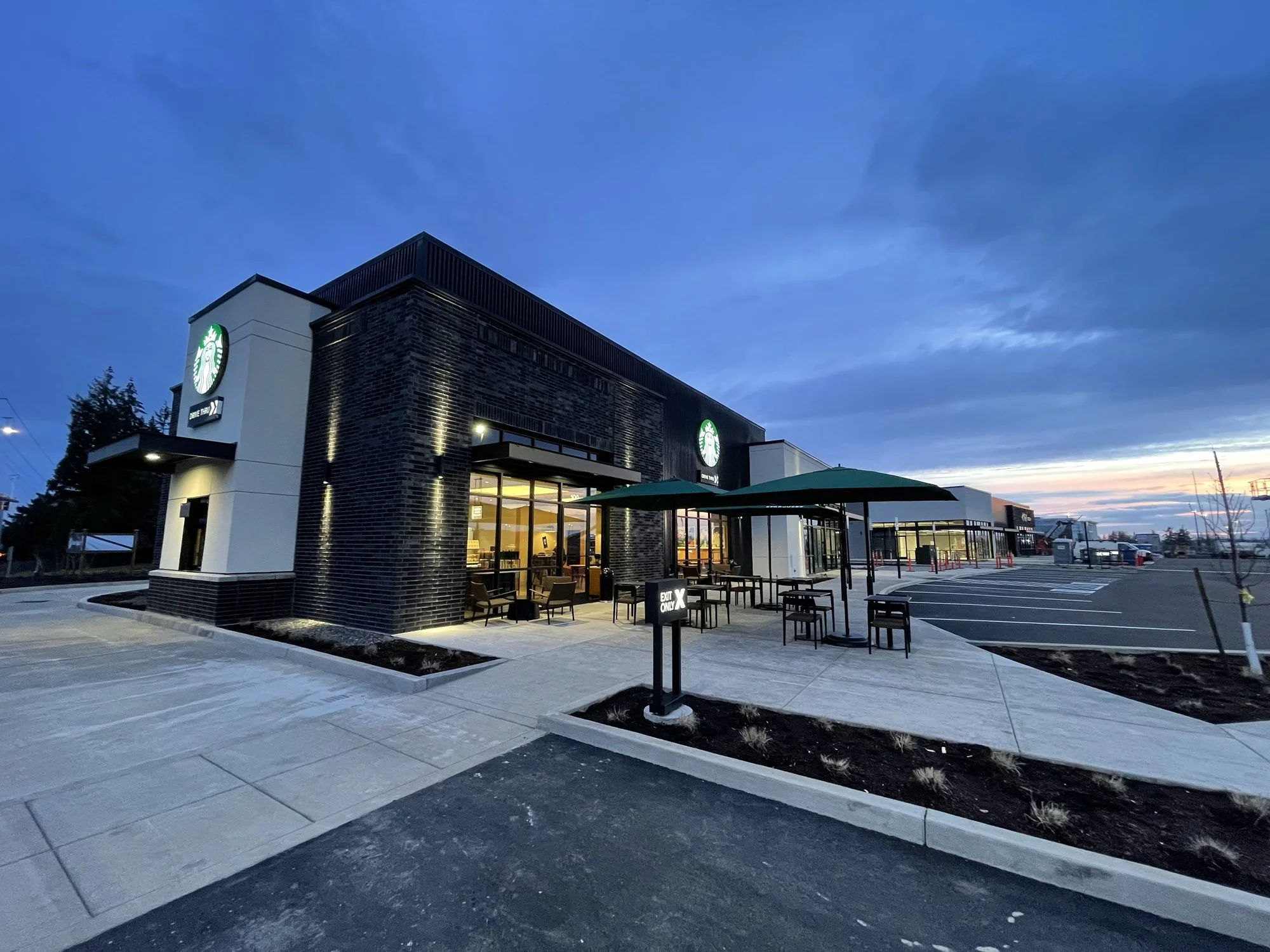 Exterior of a Starbucks coffee shop during dusk with a parking lot and outdoor seating area with tables and umbrellas.