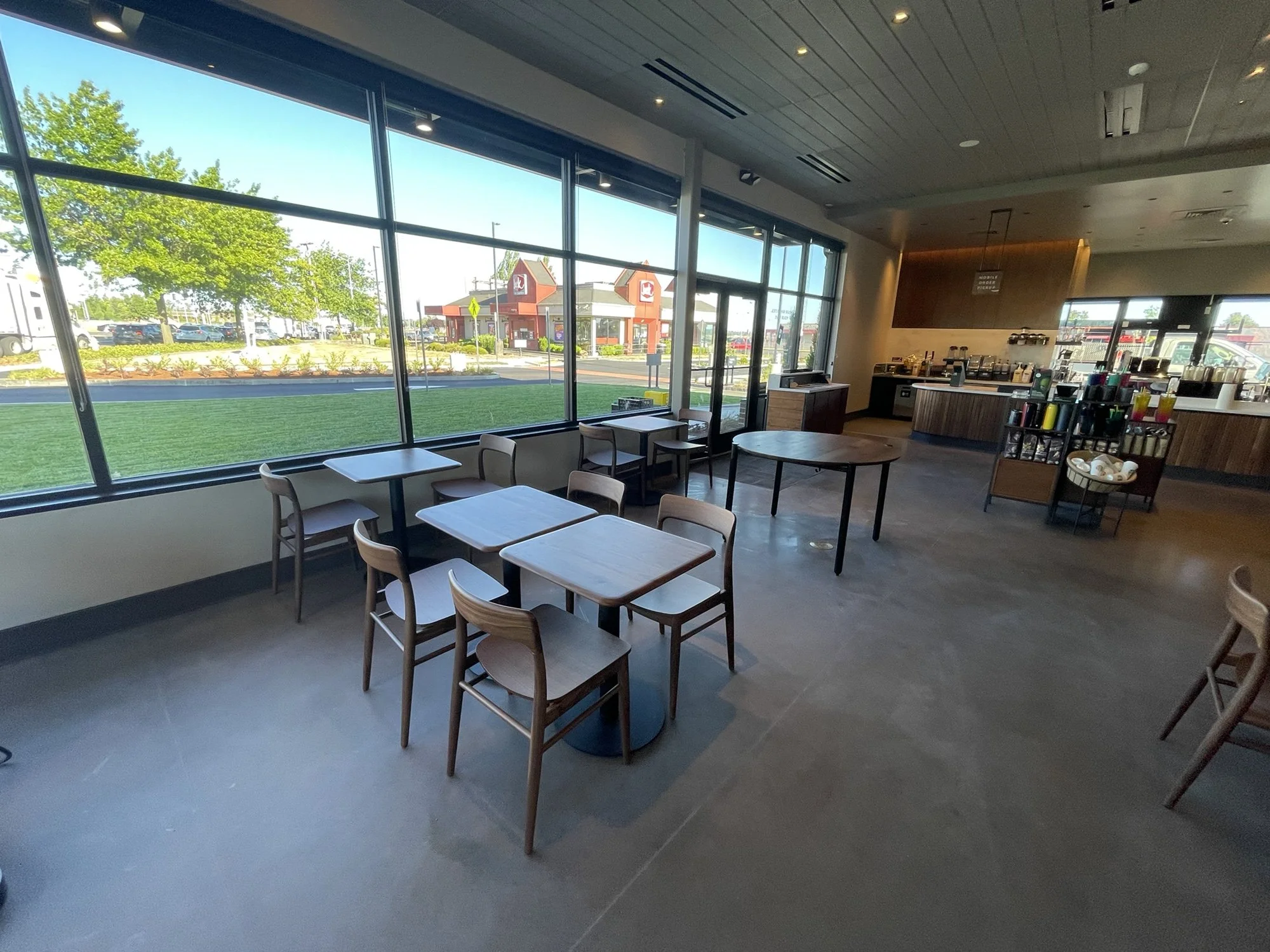 Empty indoor coffee shop or cafe with wooden tables and chairs, large window views of the parking lot and fast food restaurants outside, and a beverage station with drinks and supplies in the corner.