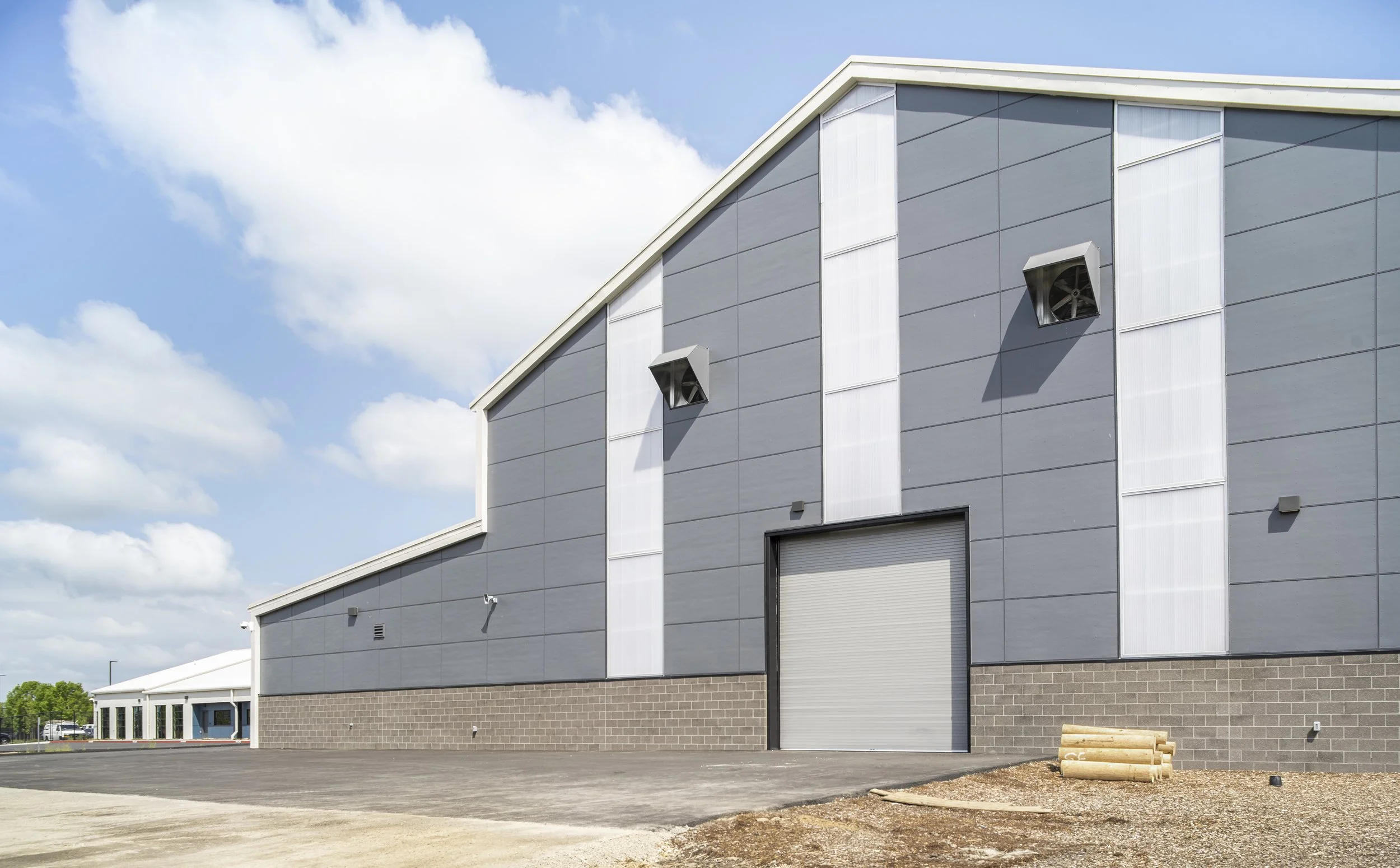Exterior view of a large, modern industrial warehouse with gray paneling, a roller door, and two wall-mounted speakers, under a partly cloudy sky.