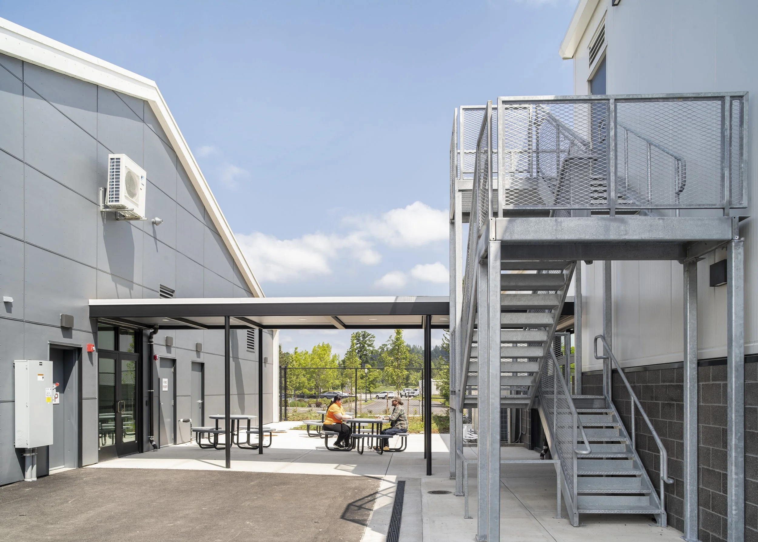 Outdoor view of a modern building with metal exterior walls, a staircase, and a shaded seating area with two people sitting at a picnic table. There are trees and parked cars in the background.