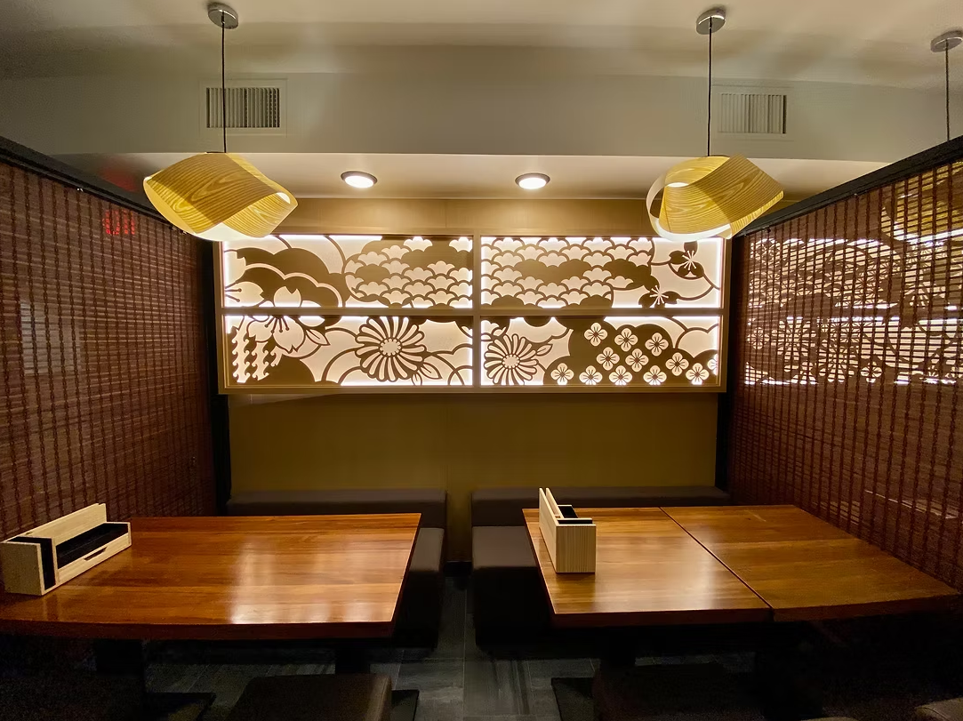 Interior of a restaurant booth with wooden tables, black cushioned seats, decorative wooden wall panels, and a floral-patterned backlit screen, with yellow hanging pendant lights.