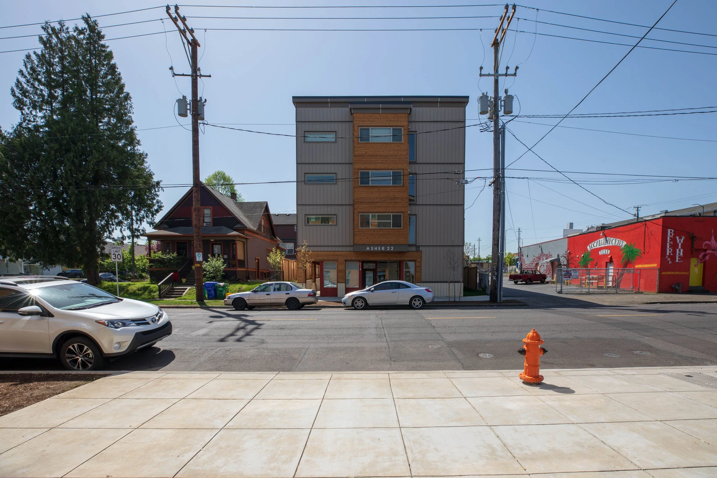 Street view with parked cars in front of a multi-story building, a red-painted building with palm trees and signs, utility poles, and a fire hydrant, under a clear blue sky.