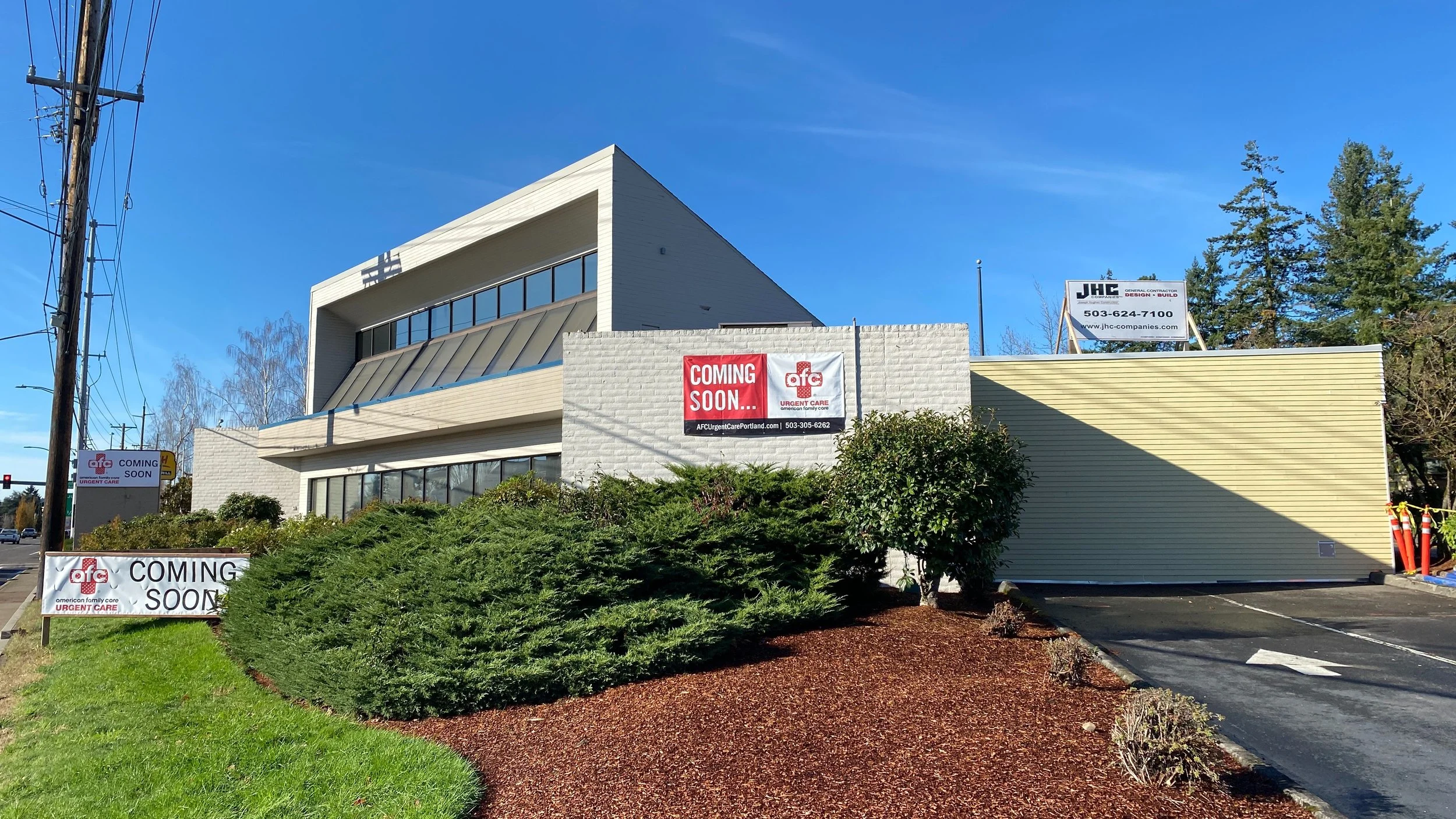 A commercial building under construction with signage indicating upcoming urgent care facility and real estate development. The building is white with large glass windows, and there are decorative shrubs and trees in front. Clear blue sky and power l