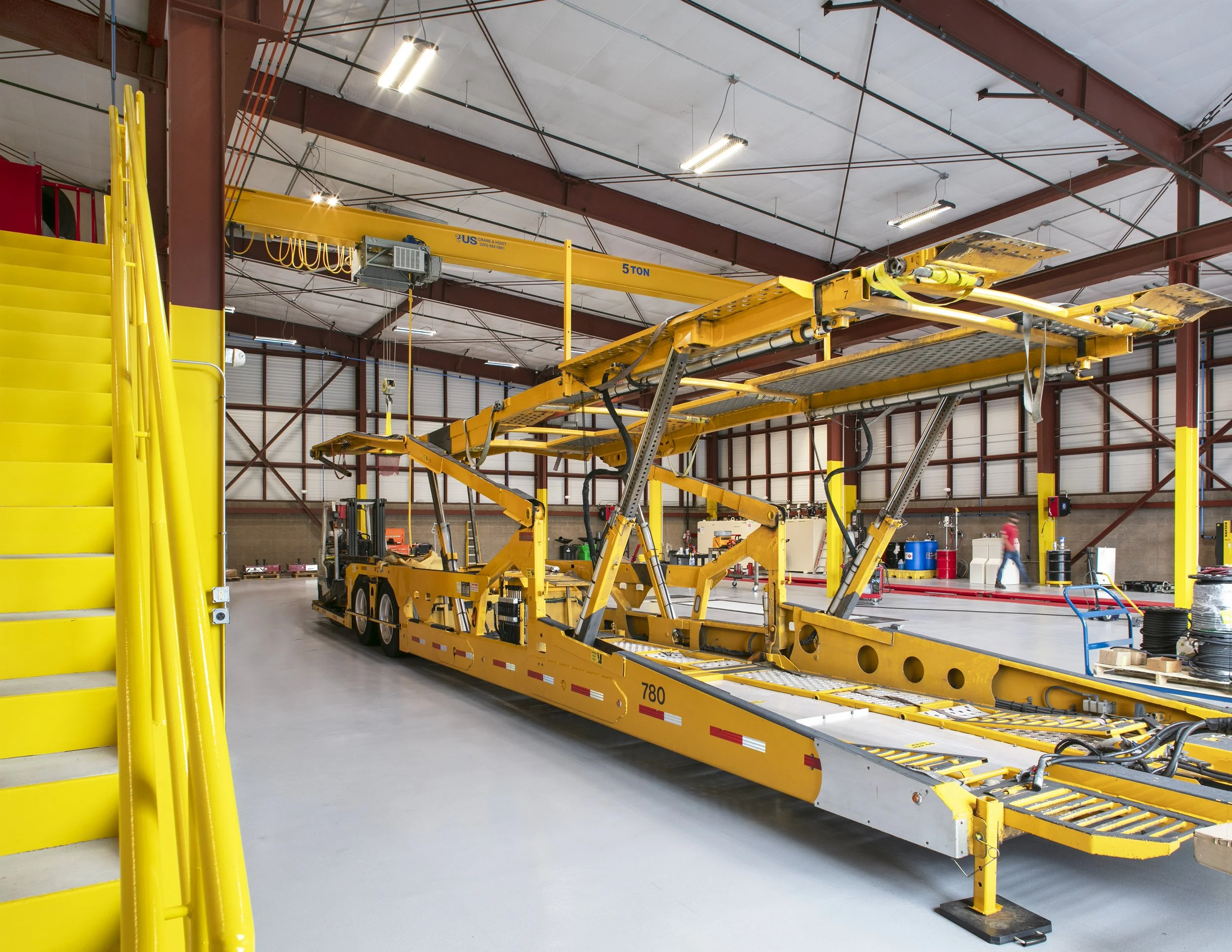 Industrial warehouse with yellow transportation equipment and staircase, overhead crane, and worker in the background.