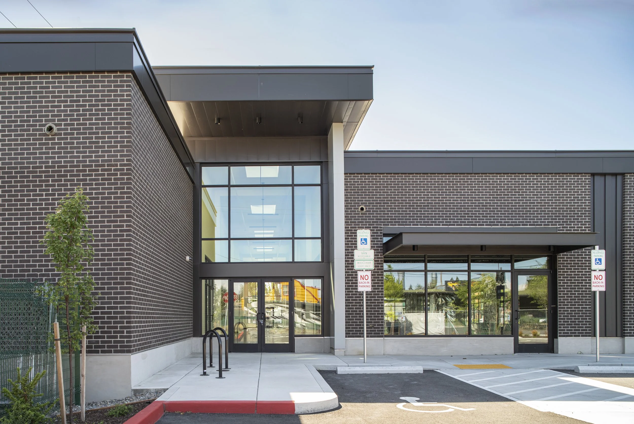 Modern building with dark brick and large glass windows, designated handicapped parking spaces, and signage, with a small landscaped area on the left.